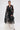 A woman stands against a white background in a romantic, rebellious look, wearing the IN BLOOM SHEER TRENCH IN BLACK with embroidered floral lace over white wide-legged pants. She accessorizes with a black gold-handle handbag and gold hoop earrings.