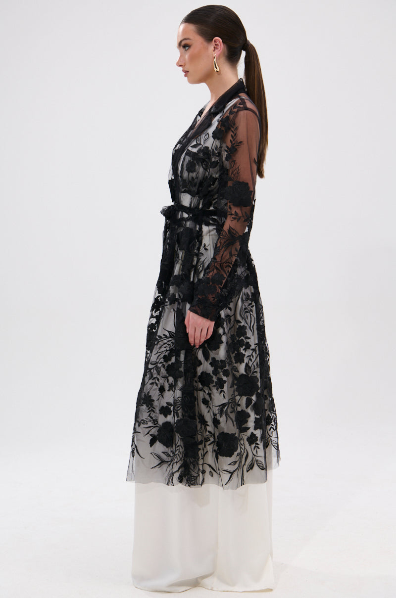 A woman in profile wears the IN BLOOM SHEER TRENCH IN BLACK, embroidered with black florals over a white dress. She has her hair in a ponytail, large hoop earrings, and stands against a plain white background for a romantic rebellious look.