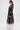 A woman in profile wears the IN BLOOM SHEER TRENCH IN BLACK, embroidered with black florals over a white dress. She has her hair in a ponytail, large hoop earrings, and stands against a plain white background for a romantic rebellious look.