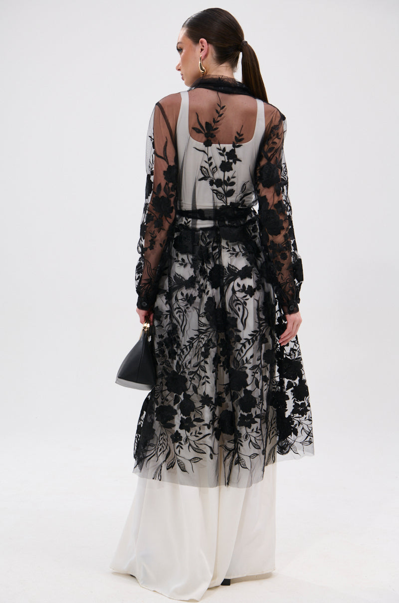 A woman with a ponytail is seen from behind wearing the IN BLOOM SHEER TRENCH IN BLACK—embroidered with floral designs—over a white dress. She carries a black handbag, embodying romantic rebellion against a plain white background.