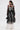 A woman with a ponytail is seen from behind wearing the IN BLOOM SHEER TRENCH IN BLACK—embroidered with floral designs—over a white dress. She carries a black handbag, embodying romantic rebellion against a plain white background.