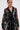 A woman with slicked-back hair and gold hoop earrings models the IN BLOOM SHEER TRENCH IN BLACK—a romantic, rebellious, sheer black trench with embroidered floral lace and a tie waist—posing confidently against a plain white background.