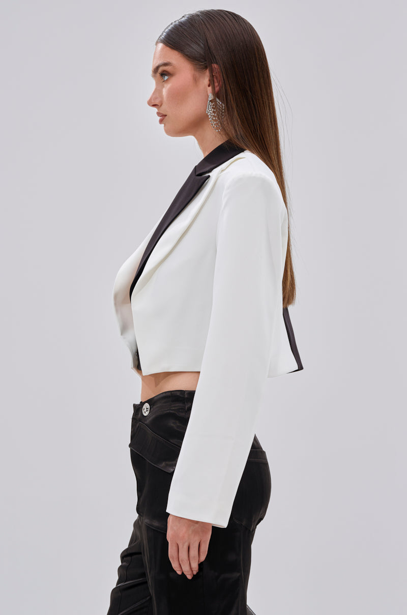 A woman with straight brown hair wears the MADE YOU LOOK DOUBLE LAYER BLAZER—a tailored white cropped jacket with black lapels—paired with black pants and statement earrings, standing in profile against a plain light background.