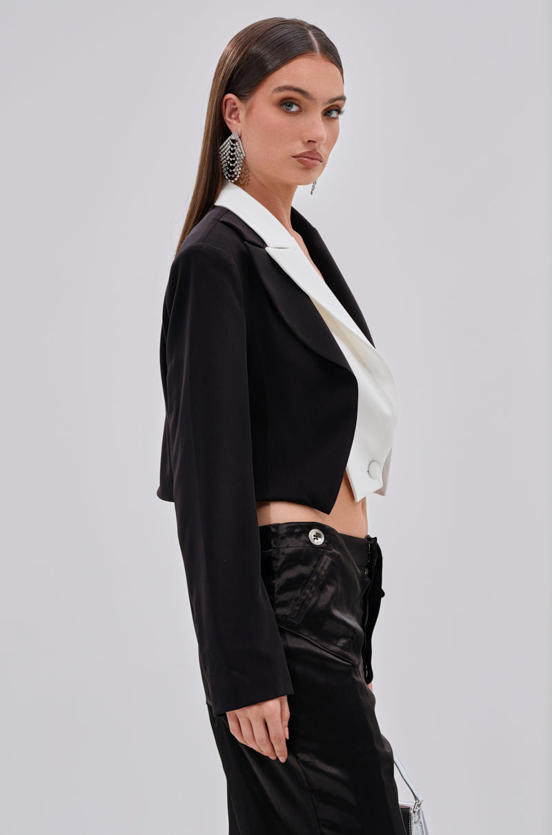 A woman with long straight brown hair wears the MADE YOU LOOK DOUBLE LAYER BLAZER—a black colorblock cropped blazer with a white lapel—paired with high-waisted black pants and statement earrings, standing sideways and looking toward the camera.