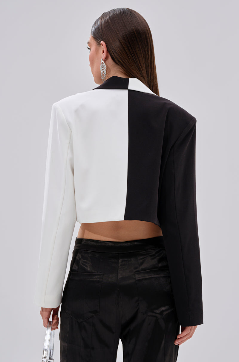 A woman faces away, wearing the MADE YOU LOOK DOUBLE LAYER BLAZER in a black and white colorblock style with black satin pants. Her straight hair is loose, and she holds a metallic purse against a plain light background.