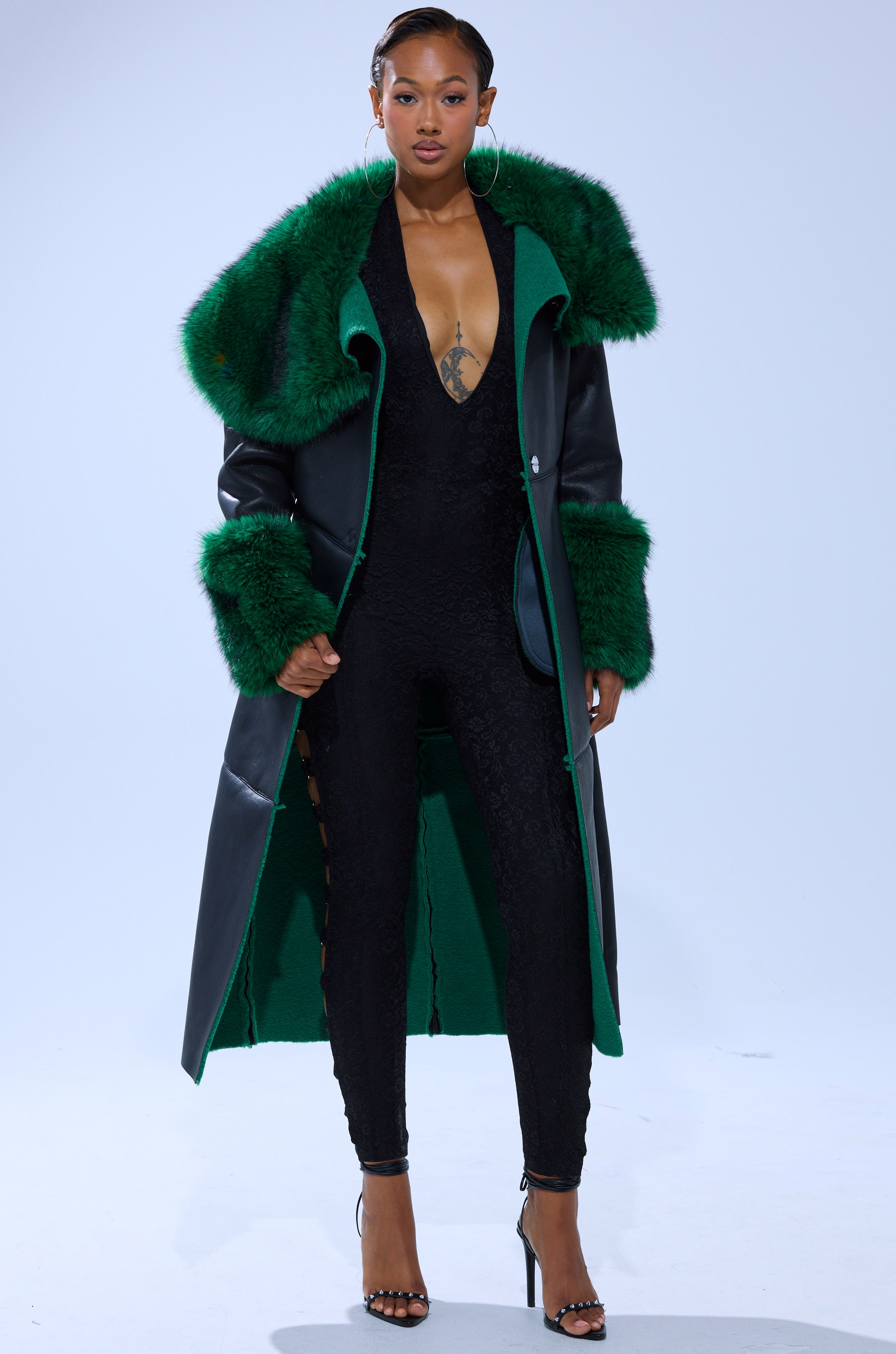 A woman stands confidently in the SEEING GREEN FUR TRIM TRENCH, a longline deep green and black coat with emerald fur trim, layered over a black lace jumpsuit and high-heeled sandals against a plain light background.
