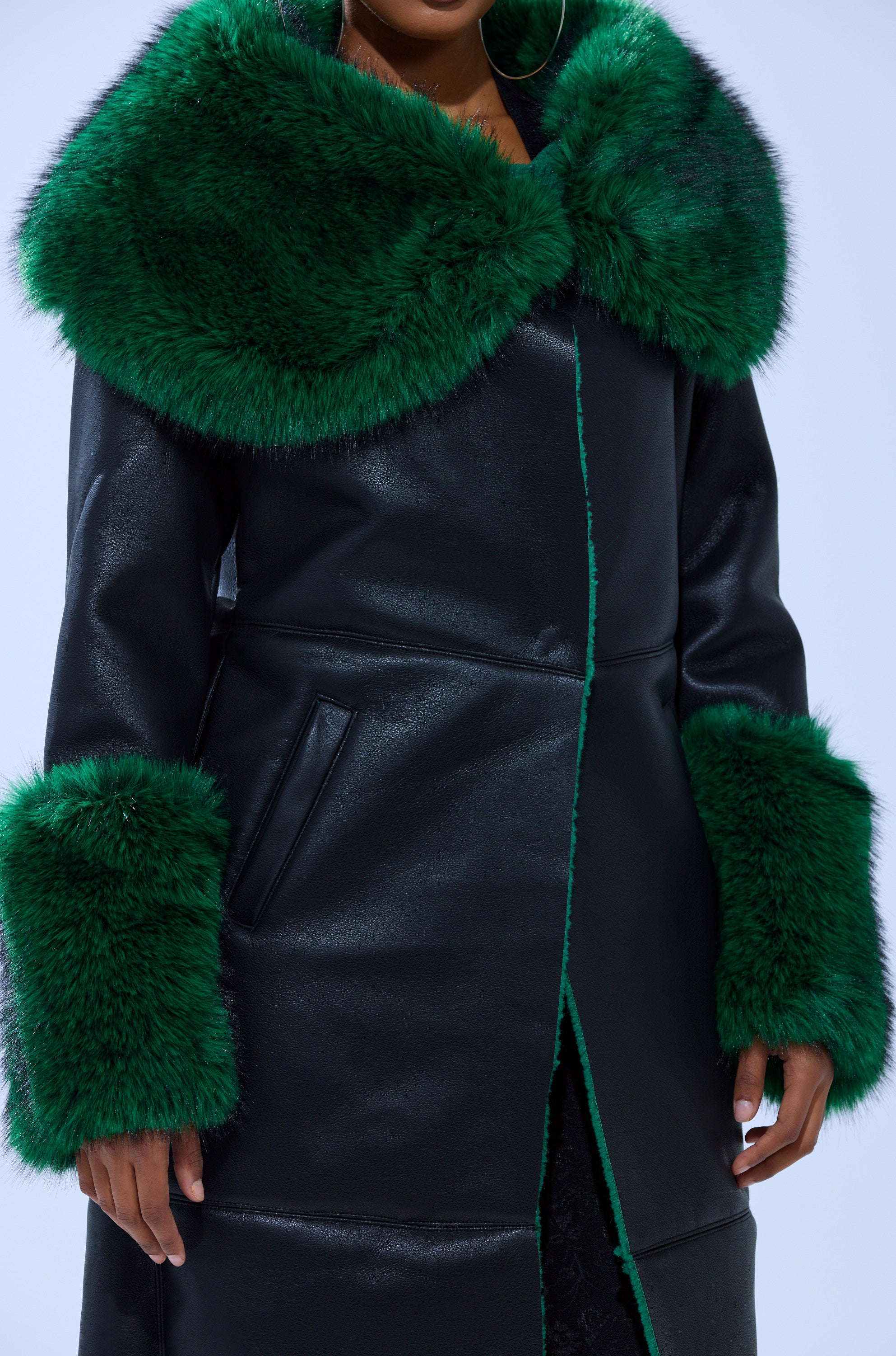 Person in the SEEING GREEN FUR TRIM TRENCH—a black faux leather jacket with oversized emerald green faux fur collar and cuffs, side pockets—stands against a plain background, face partially out of frame.