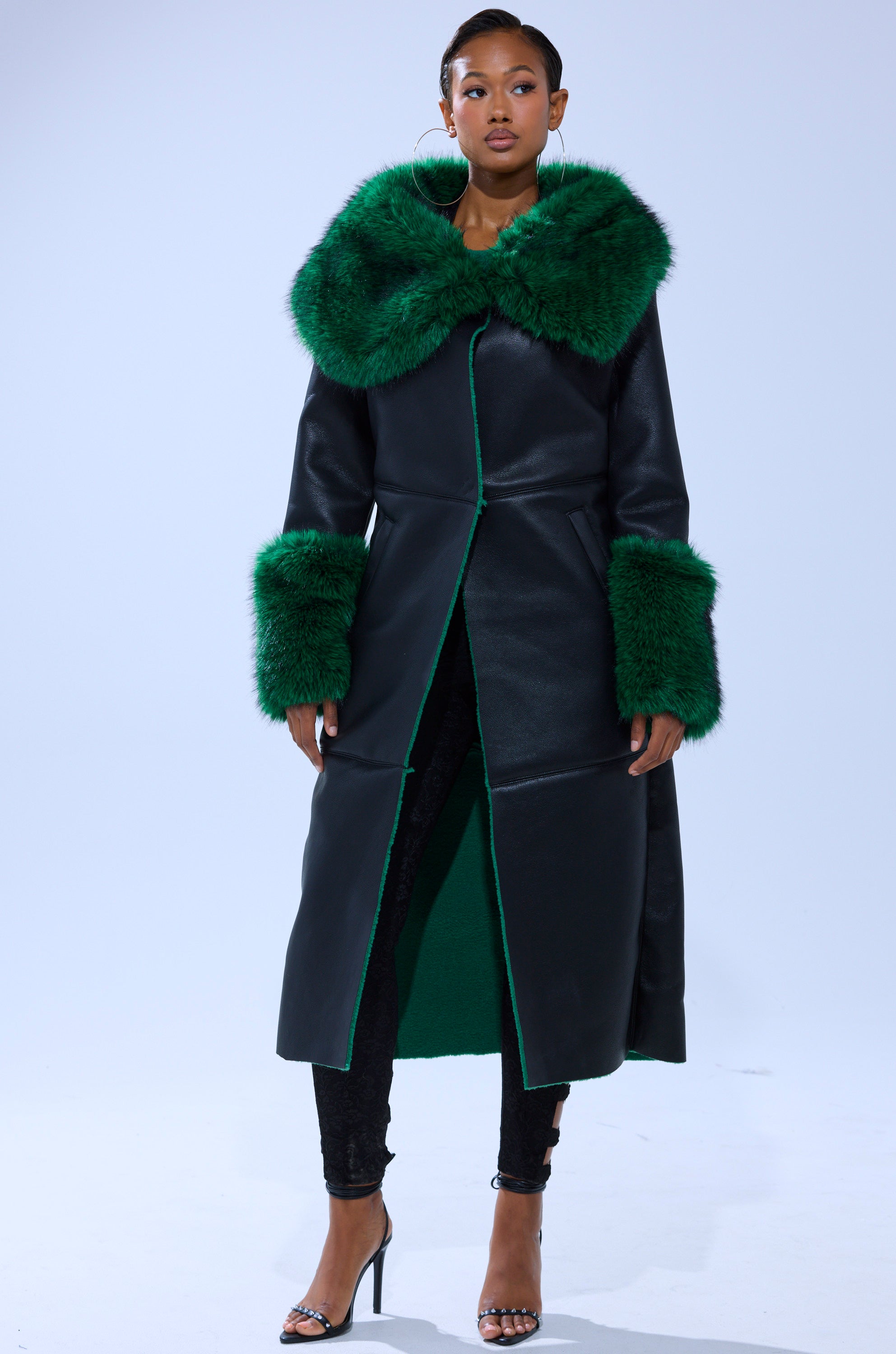 A woman models the SEEING GREEN FUR TRIM TRENCH—a black faux leather coat with emerald green fur trim—paired with black pants and high-heeled sandals, posing against a light, plain background.