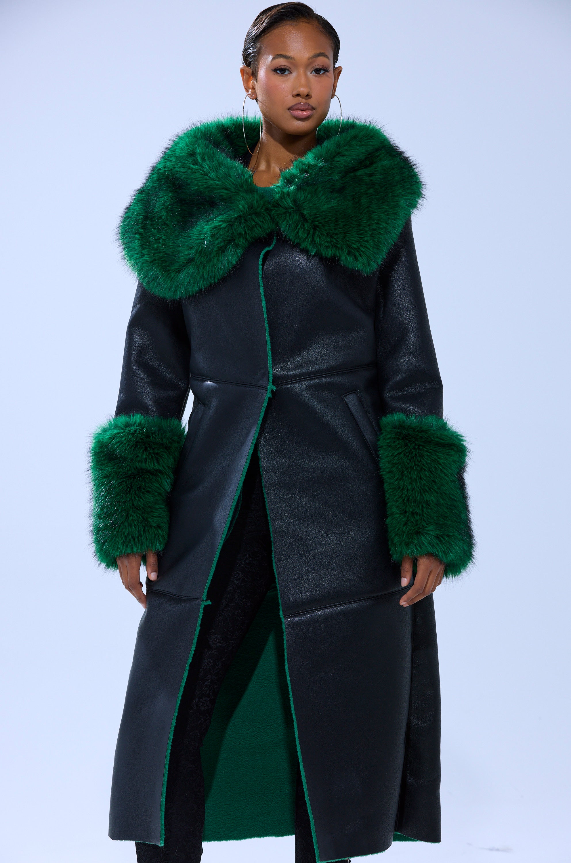 A woman models the SEEING GREEN FUR TRIM TRENCH—a black longline coat featuring bold emerald green fur on the collar and cuffs—standing against a plain light background.
