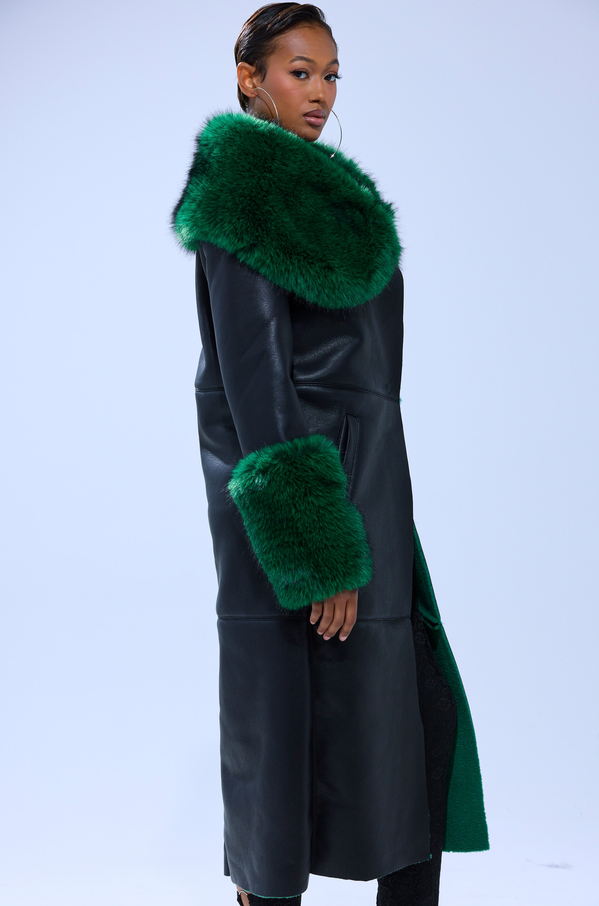 A woman poses against a plain light background wearing the SEEING GREEN FUR TRIM TRENCH, a longline faux leather coat accented with bold emerald green fur on the collar and cuffs.