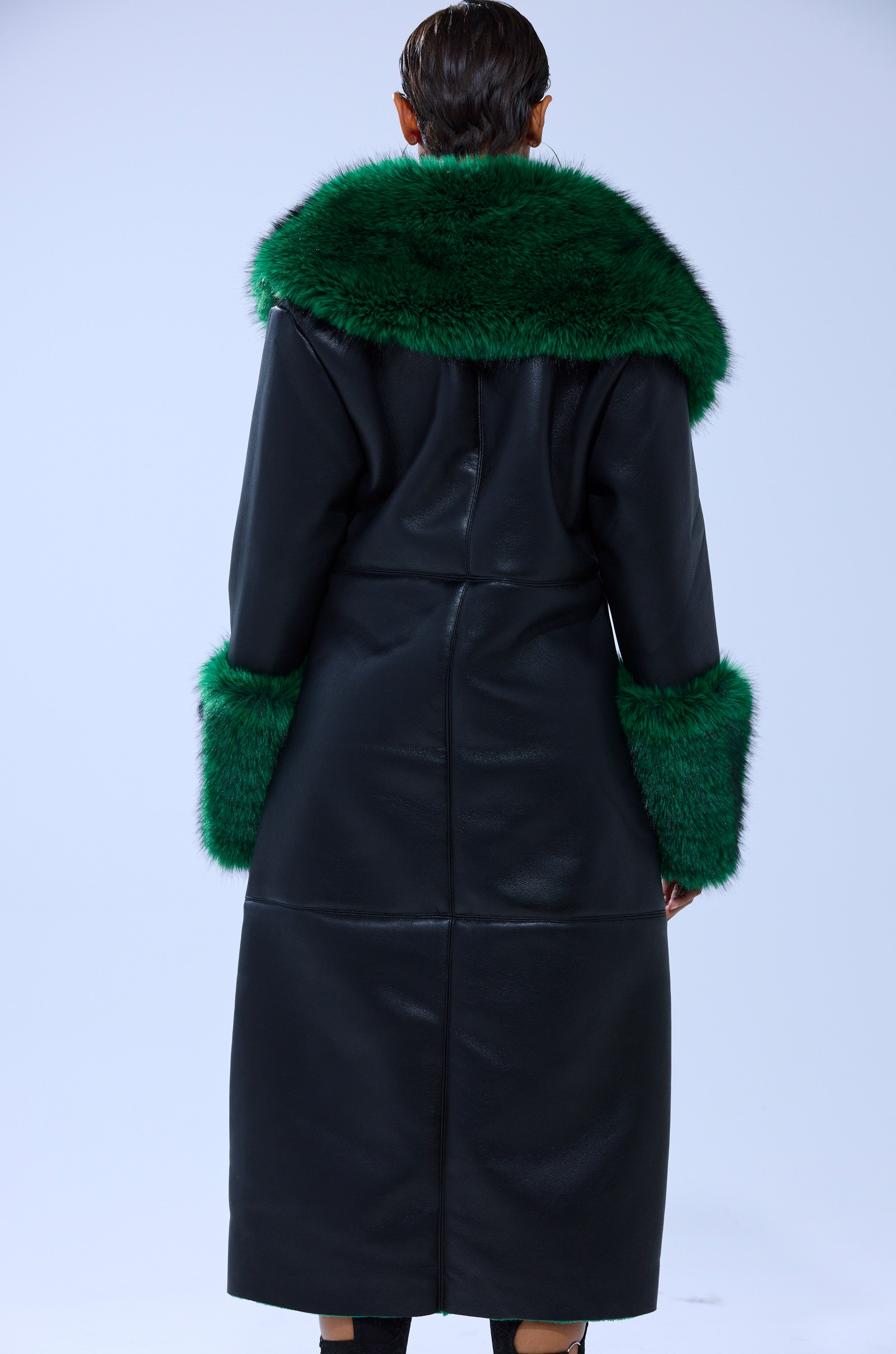 Someone wearing the SEEING GREEN FUR TRIM TRENCH— a long black faux leather coat with bold emerald green fur trim on the collar and cuffs—stands with their back to the camera against a plain light background.