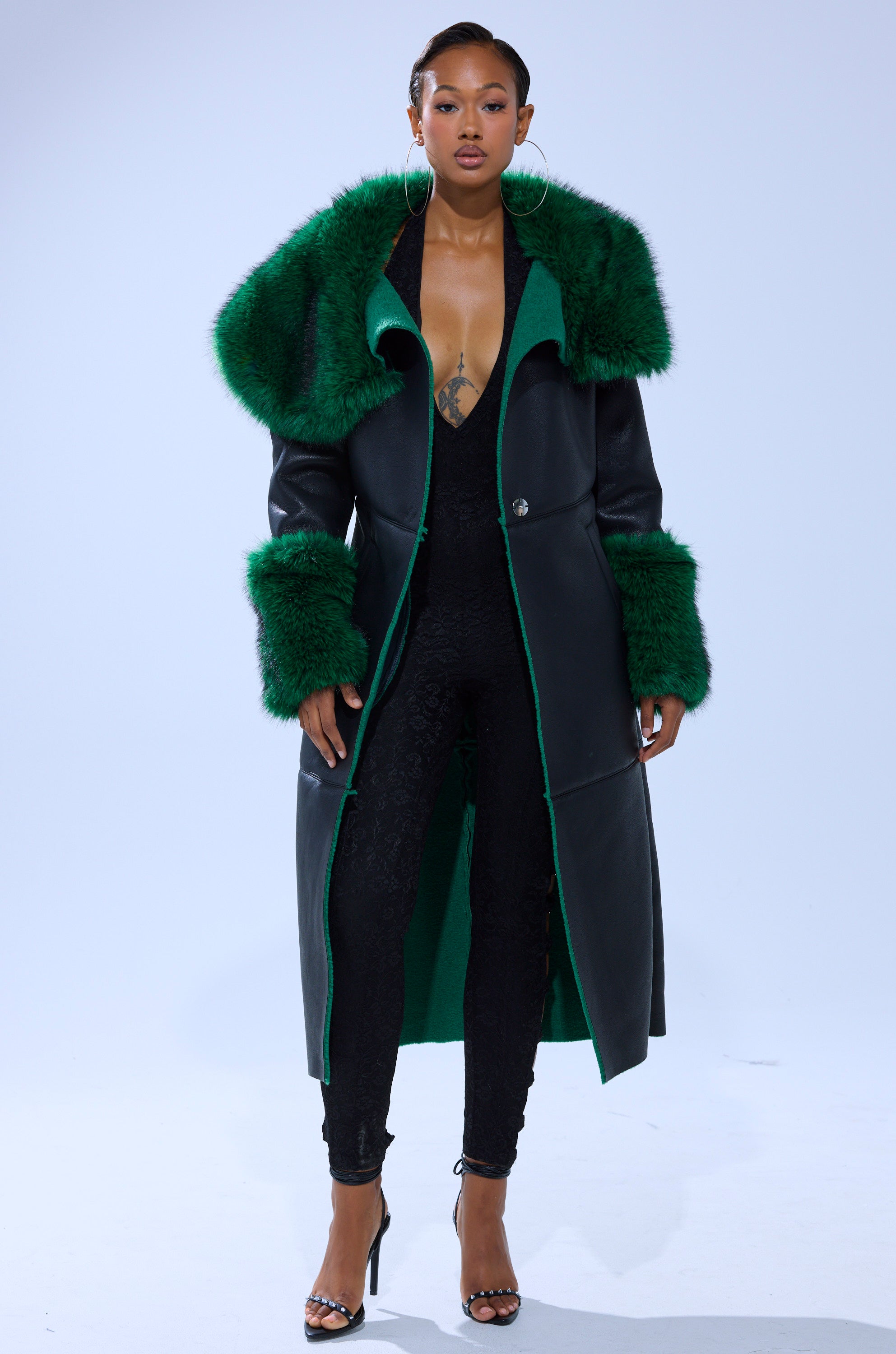 A woman models the SEEING GREEN FUR TRIM TRENCH, featuring a bold emerald collar and cuffs, over a black lace jumpsuit with open-toed heels against a plain light background.