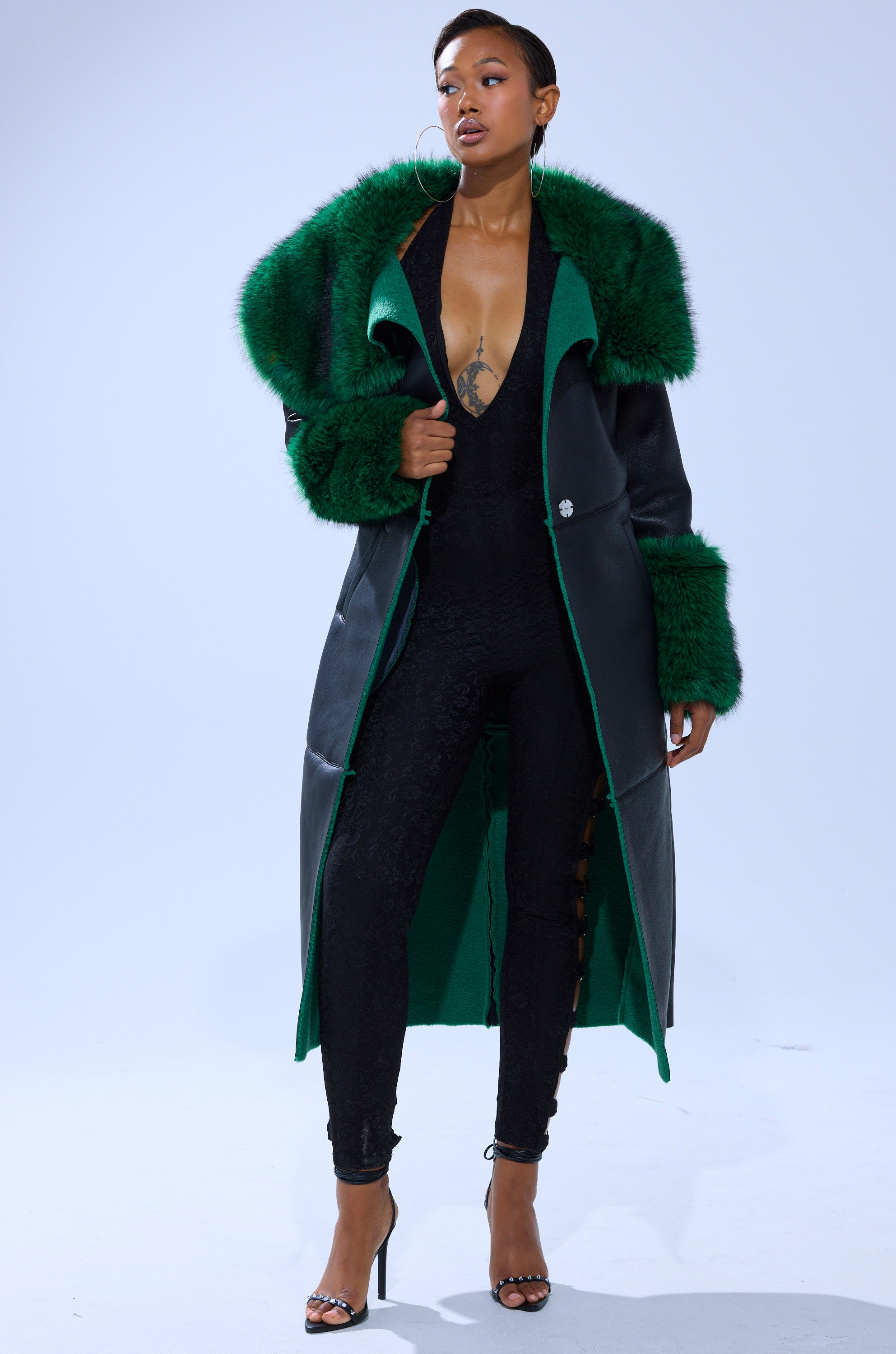 A woman poses confidently in the SEEING GREEN FUR TRIM TRENCH over a fitted black jumpsuit and high heels, standing against a plain light background.
