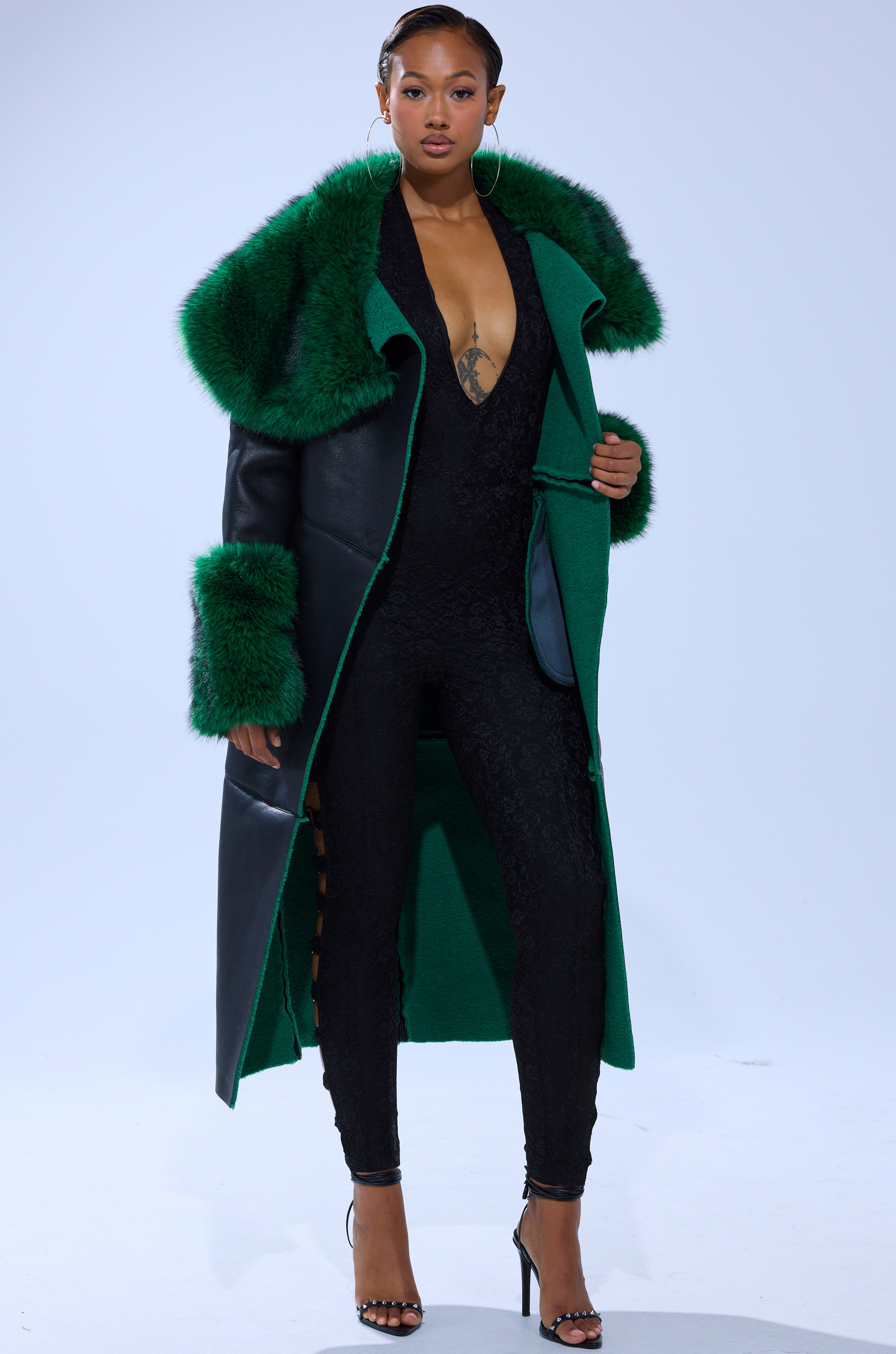 A woman stands confidently in black heels, wearing the SEEING GREEN FUR TRIM TRENCH over a black lace jumpsuit with a plunging neckline and lace-up sides, set against a plain light background.