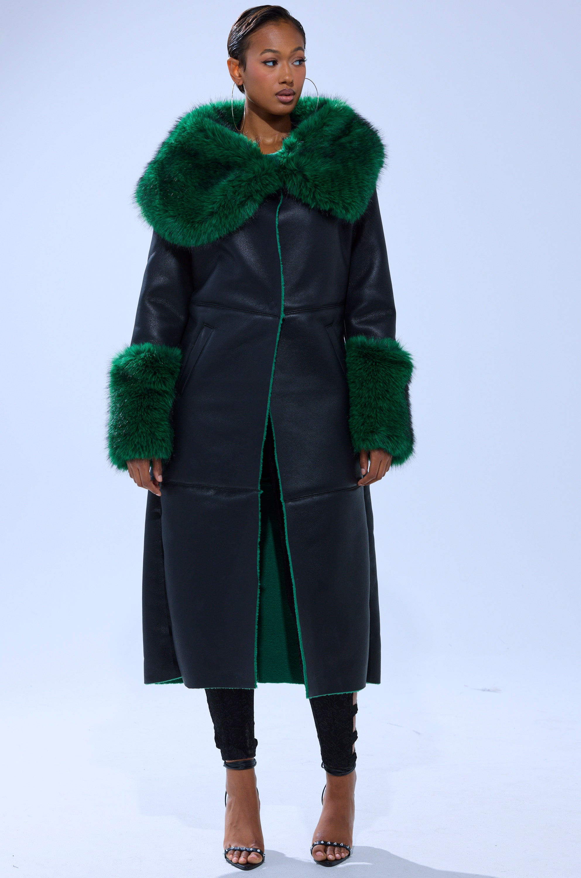 A woman models the SEEING GREEN FUR TRIM TRENCH—a long black faux leather coat with oversized green faux fur collar and cuffs—paired with black pants and open-toed heels, posing against a light background.