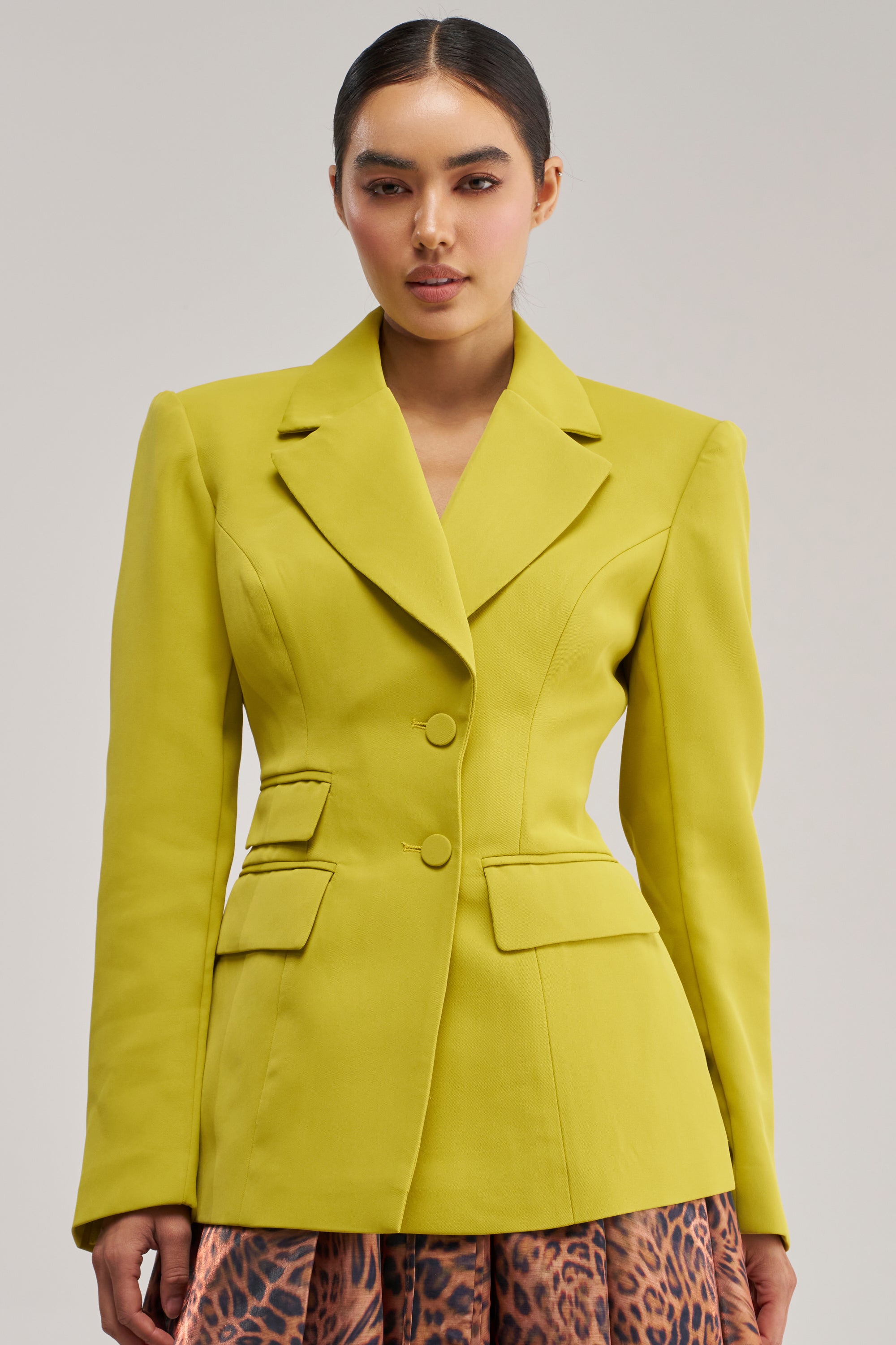 A woman stands against a plain light background wearing THE BLUEPRINT BLAZER IN CHARTREUSE—a bold, fitted suit jacket featuring a two-button closure and flap pockets—paired with a leopard print skirt. This structured blazer creates an eye-catching statement in bold officewear.