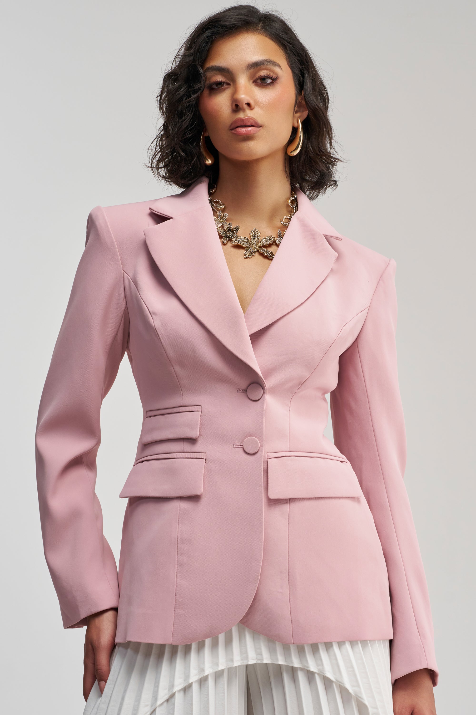 Woman with wavy dark hair wearing THE BLUEPRINT BLAZER IN MAUVE, paired with white pleated pants, a gold necklace, and gold hoop earrings, standing against a plain light background for a business-chic look.