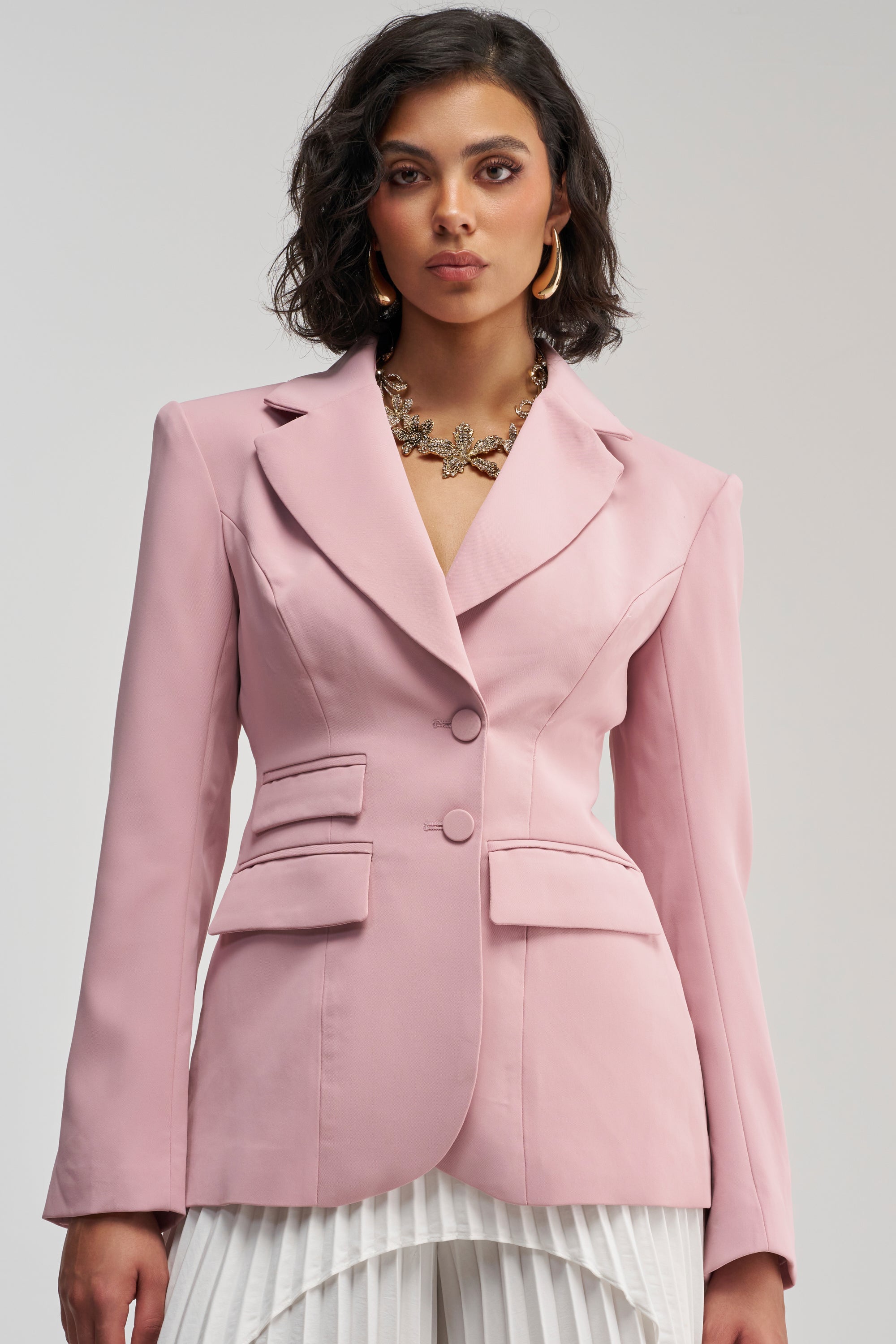 A woman models THE BLUEPRINT BLAZER IN MAUVE, featuring large lapels and double front pockets, styled with a statement necklace and white pleated pants against a simple backdrop for a polished business-chic appearance.