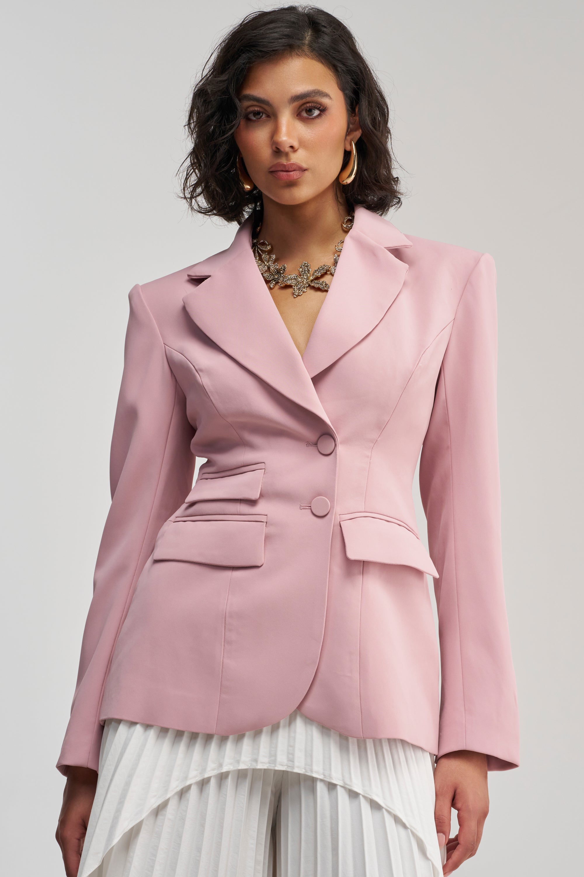 A woman with short dark hair styles THE BLUEPRINT BLAZER IN MAUVE, featuring a tailored fit and flap pockets, paired with a pleated white skirt for a business-chic ensemble. She completes the look with gold hoop earrings and a chunky necklace, standing against a plain background.