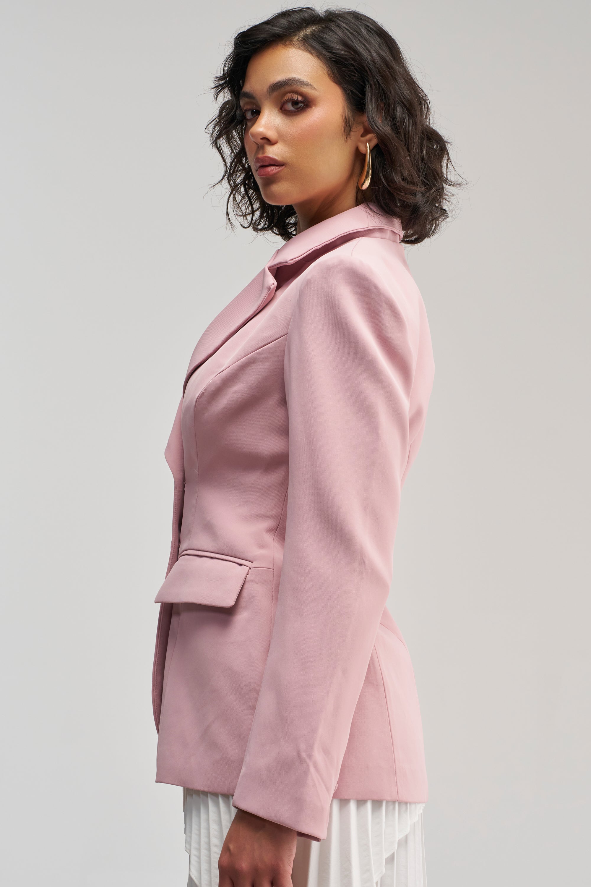 Woman with wavy dark hair wearing THE BLUEPRINT BLAZER IN MAUVE and a white skirt, standing sideways and looking confidently at the camera against a plain background.