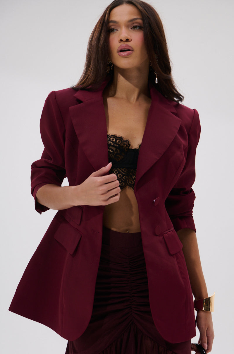 A woman stands against a plain light background, wearing THE BLUEPRINT ASYMMETRICAL BLAZER over a black lace bralette and matching skirt. She has long wavy brown hair and accessorizes with earrings and a gold cuff bracelet.