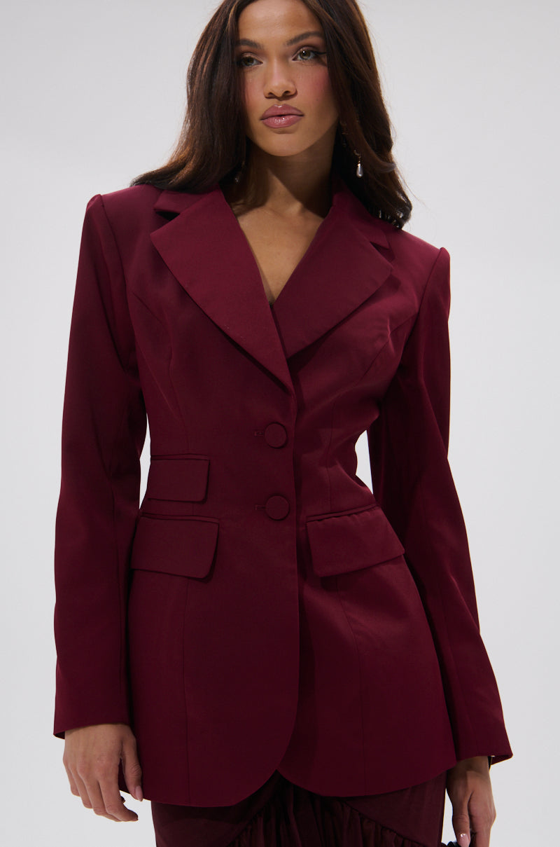 A woman with long brown hair confidently models THE BLUEPRINT ASYMMETRICAL BLAZER, featuring a structured design and wide lapels, against a plain light gray background.