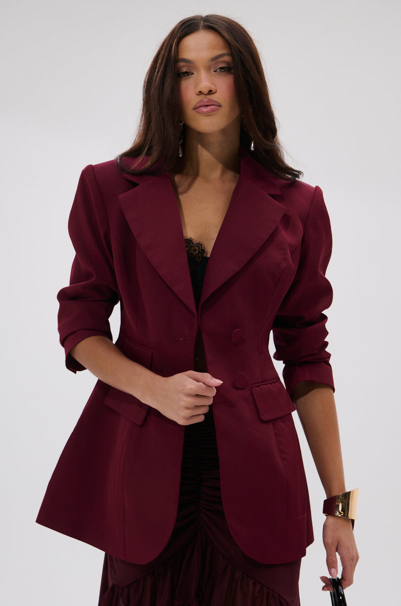 A woman with long brown hair wears THE BLUEPRINT ASYMMETRICAL BLAZER in burgundy over a black lace top and dark skirt. She accessorizes with a gold cuff bracelet and poses confidently against a plain, light background.