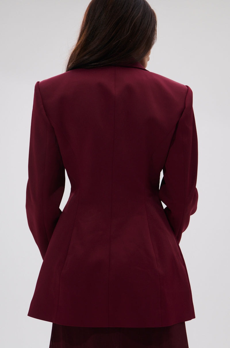 A person with long dark hair, seen from behind, wears THE BLUEPRINT ASYMMETRICAL BLAZER in maroon against a plain light background.