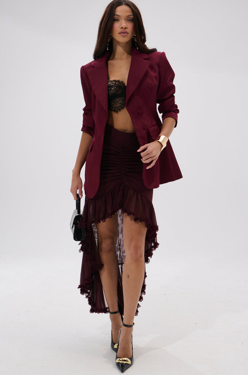 A woman walks confidently in THE BLUEPRINT ASYMMETRICAL BLAZER, styled with a black lace bralette, a high-low ruched skirt with lace trim, black heels, a cuff bracelet, and a small black handbag.
