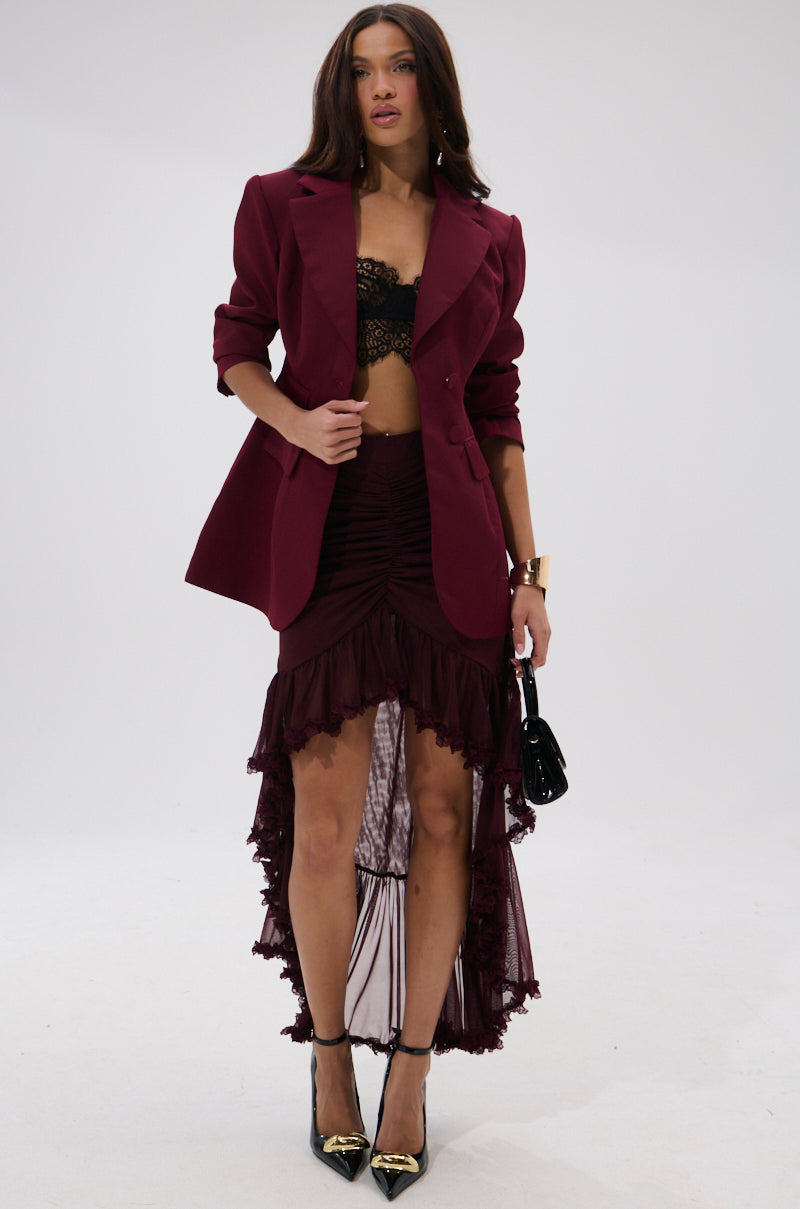 A woman models THE BLUEPRINT ASYMMETRICAL BLAZER over a black lace bralette, paired with a matching high-low ruffled skirt, black heels with gold tips, and a small black handbag against a plain white background.