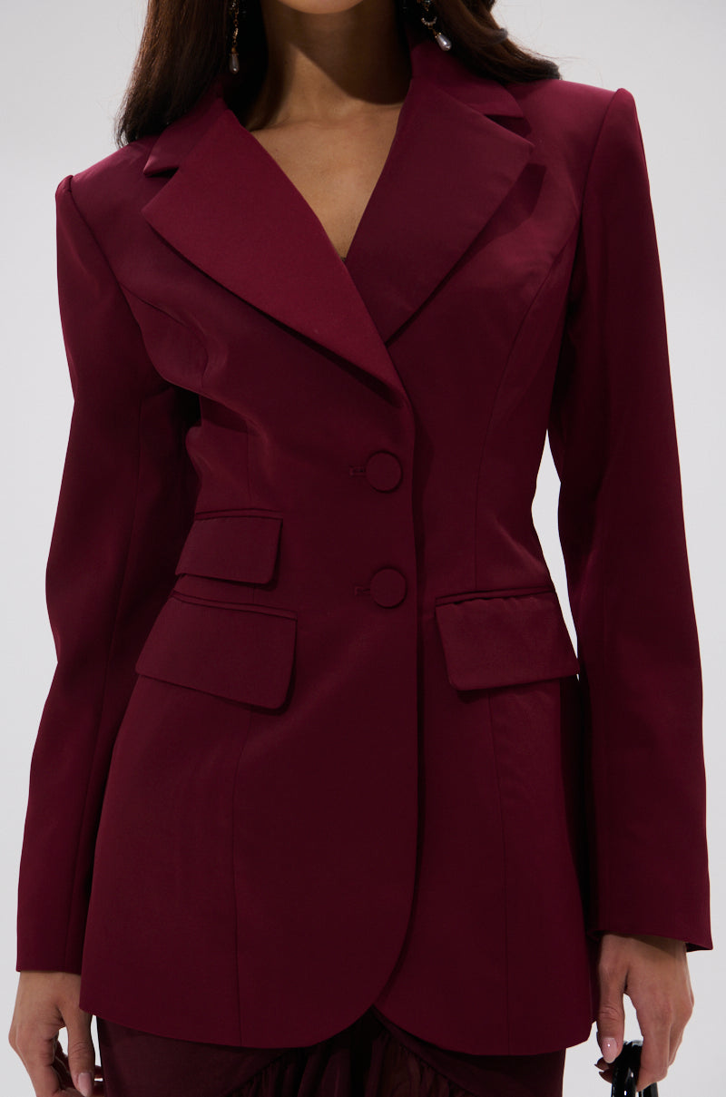 A woman wears THE BLUEPRINT ASYMMETRICAL BLAZER, a tailored deep burgundy jacket with two buttons, notched lapels, and flap pockets. She stands against a plain light background with her head partially out of the frame.