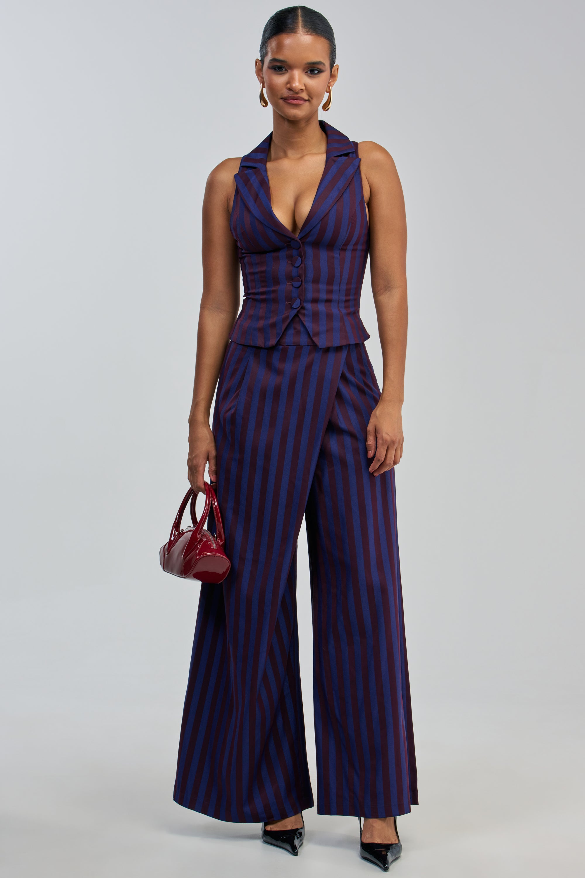 A woman wears the MANHATTAN ASYMMETRICAL STRIPE TROUSER—wide-leg pants featuring an asymmetrical blue and purple stripe design—paired with a matching sleeveless, deep V-neck vest. She accessorizes with a small red handbag and black heels, standing against a plain light background.