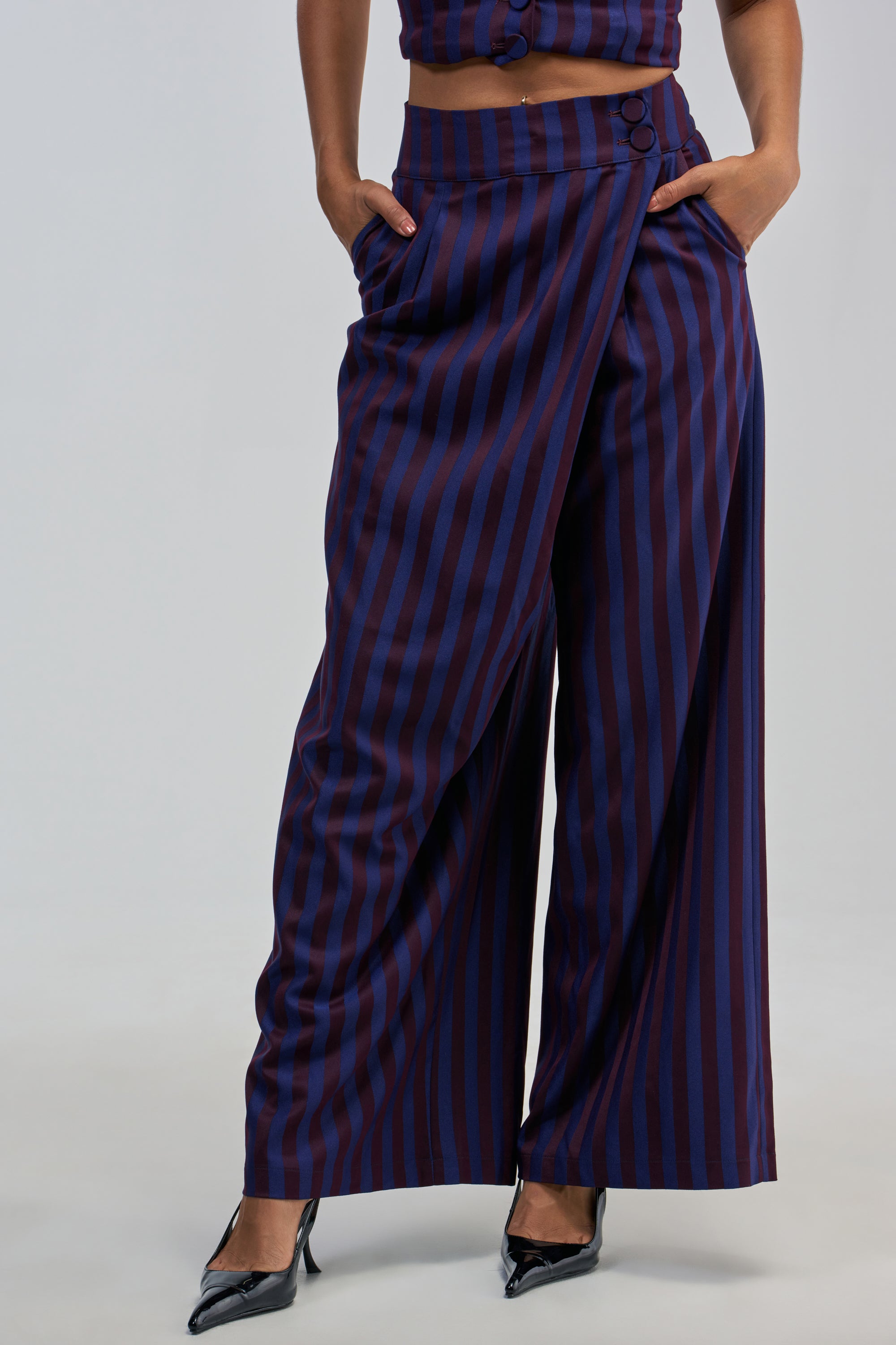 Wearing the MANHATTAN ASYMMETRICAL STRIPE TROUSER—featuring a wide-leg, high-rise fit with vertical navy and burgundy stripes—paired with a matching top and black pointed-toe heels. The person has their hands in their pockets against a plain, light-colored background.