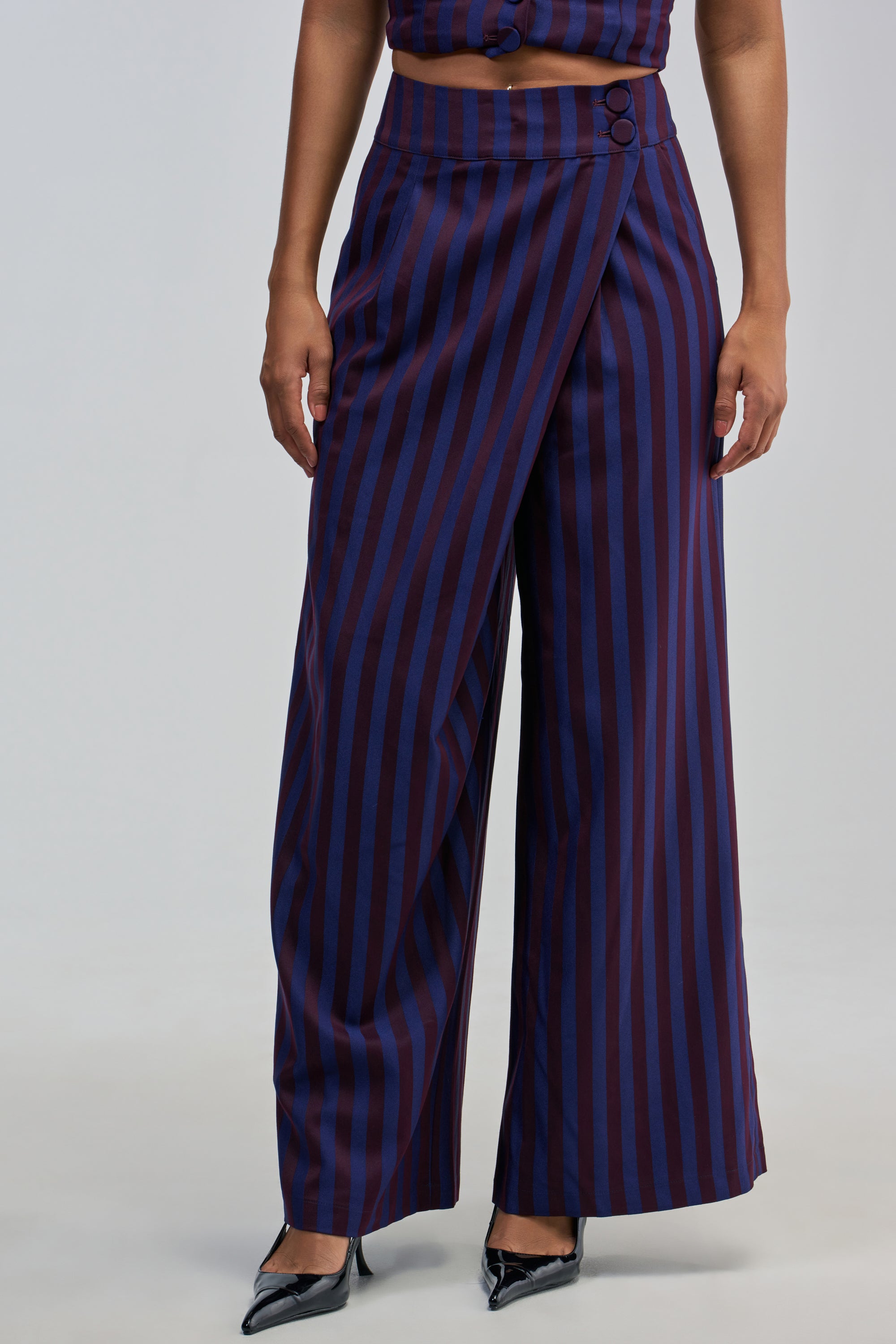 A woman is seen from the waist down wearing the MANHATTAN ASYMMETRICAL STRIPE TROUSER—high-waisted, wide-leg pants featuring vertical navy and burgundy stripes and a distinctive wrap detail with two buttons at the waist. She completes the look with black pointed-toe heels, showcasing an effortlessly chic silhouette.