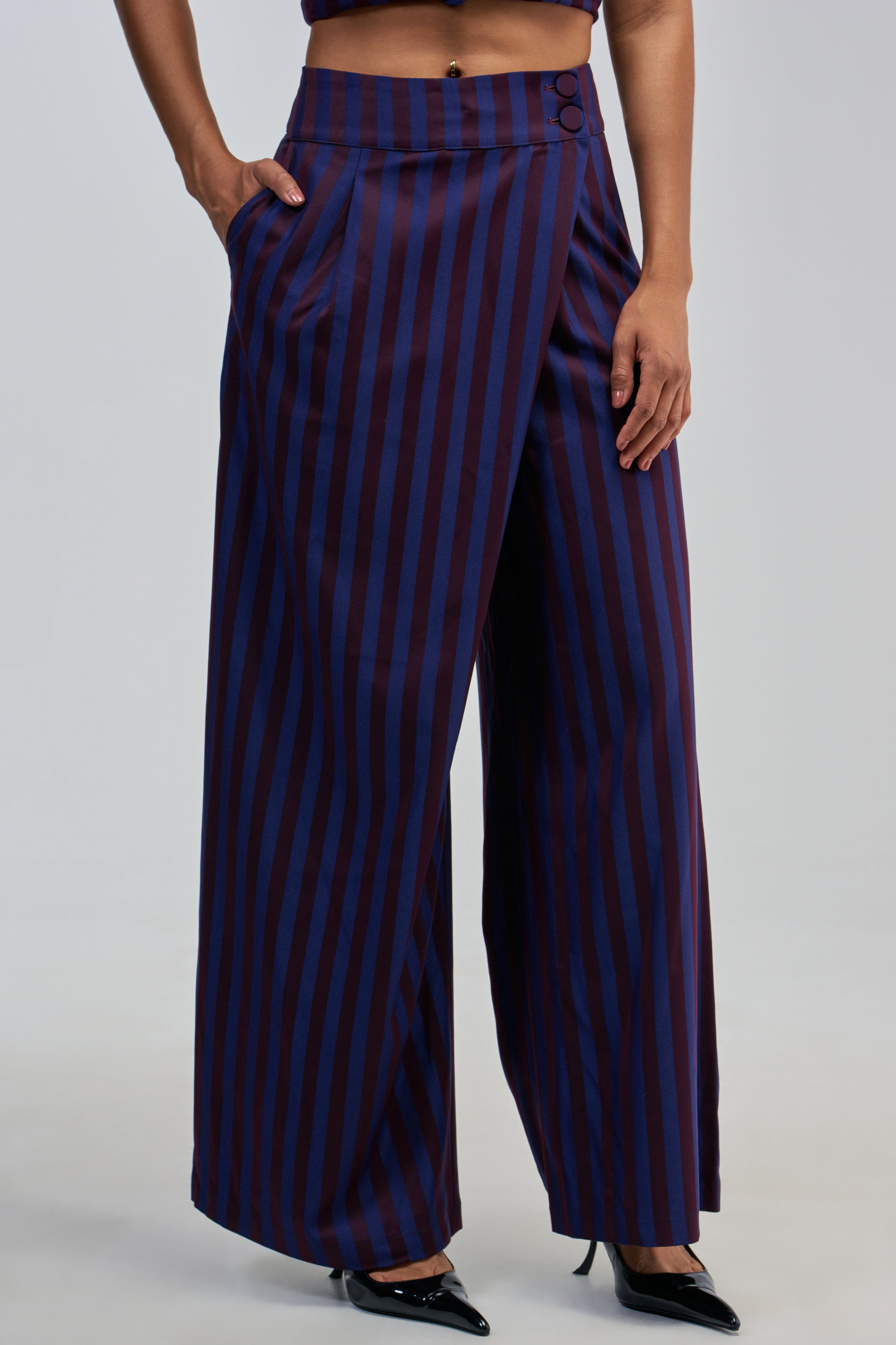 A person wearing the MANHATTAN ASYMMETRICAL STRIPE TROUSER, featuring a high-rise fit and flattering wide-leg silhouette with vertical navy and burgundy stripes, paired with black pointed-toe shoes.
