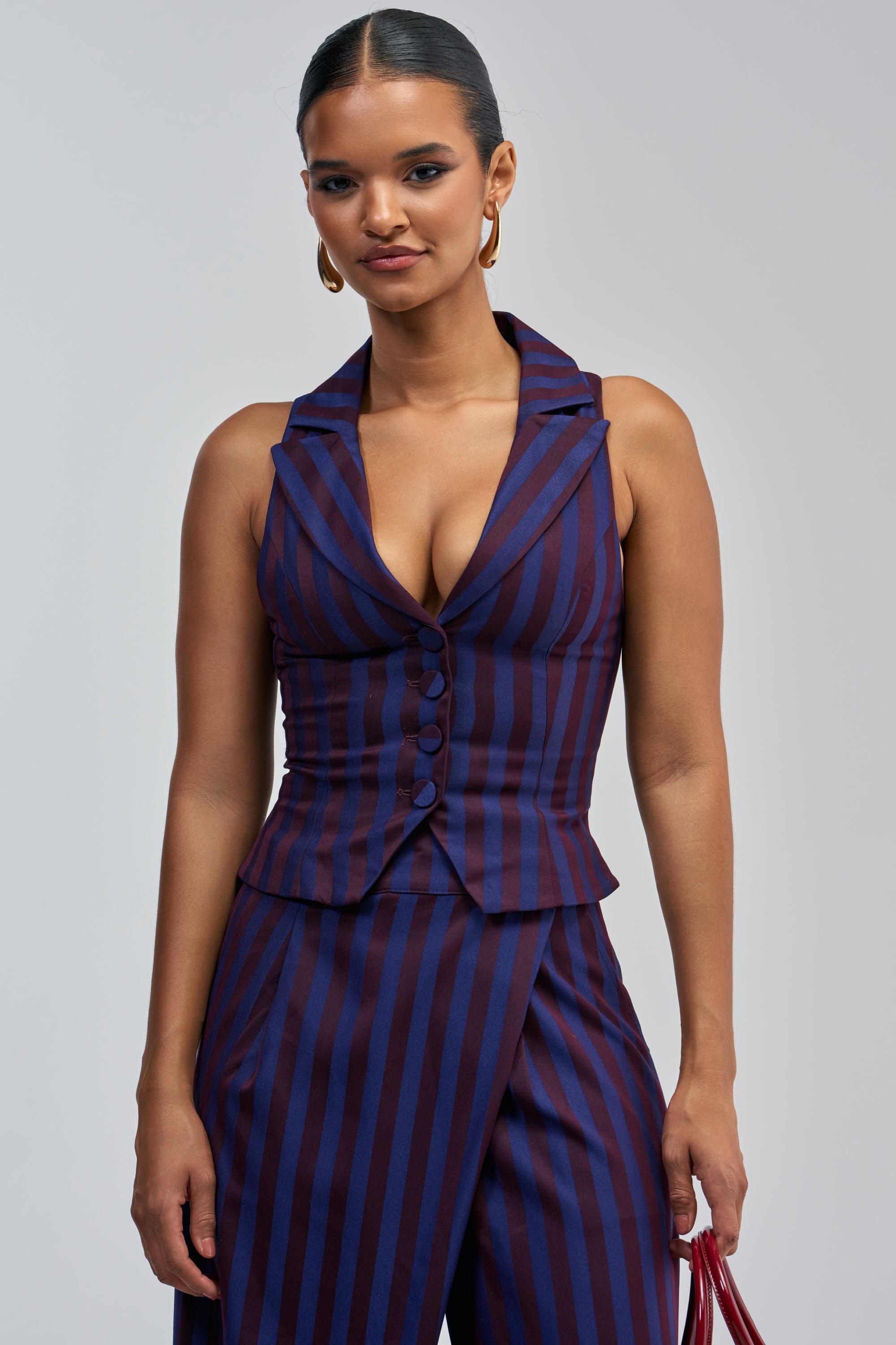 Wearing the MANHATTAN STRIPE VEST, a woman stands confidently against a plain background, her sophisticated workwear look defined by the sleeveless, deep v-neck top and wide-leg pants featuring vertical blue and purple stripes.