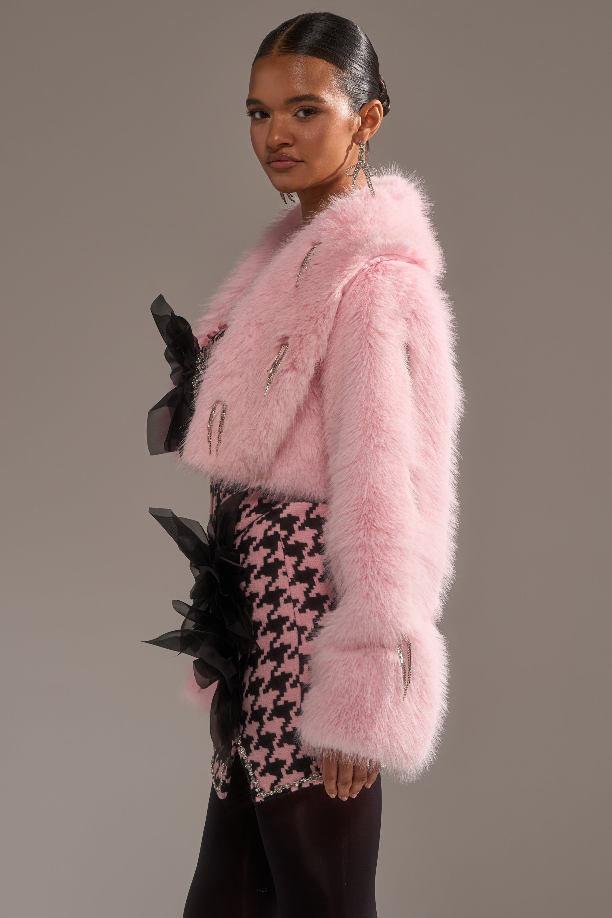 A woman poses sideways in the SWEET TALK EMBELLISHED FAUX FUR jacket with oversized sleeves, metallic accents, a black-and-pink houndstooth skirt, black tights, and statement earrings. 4