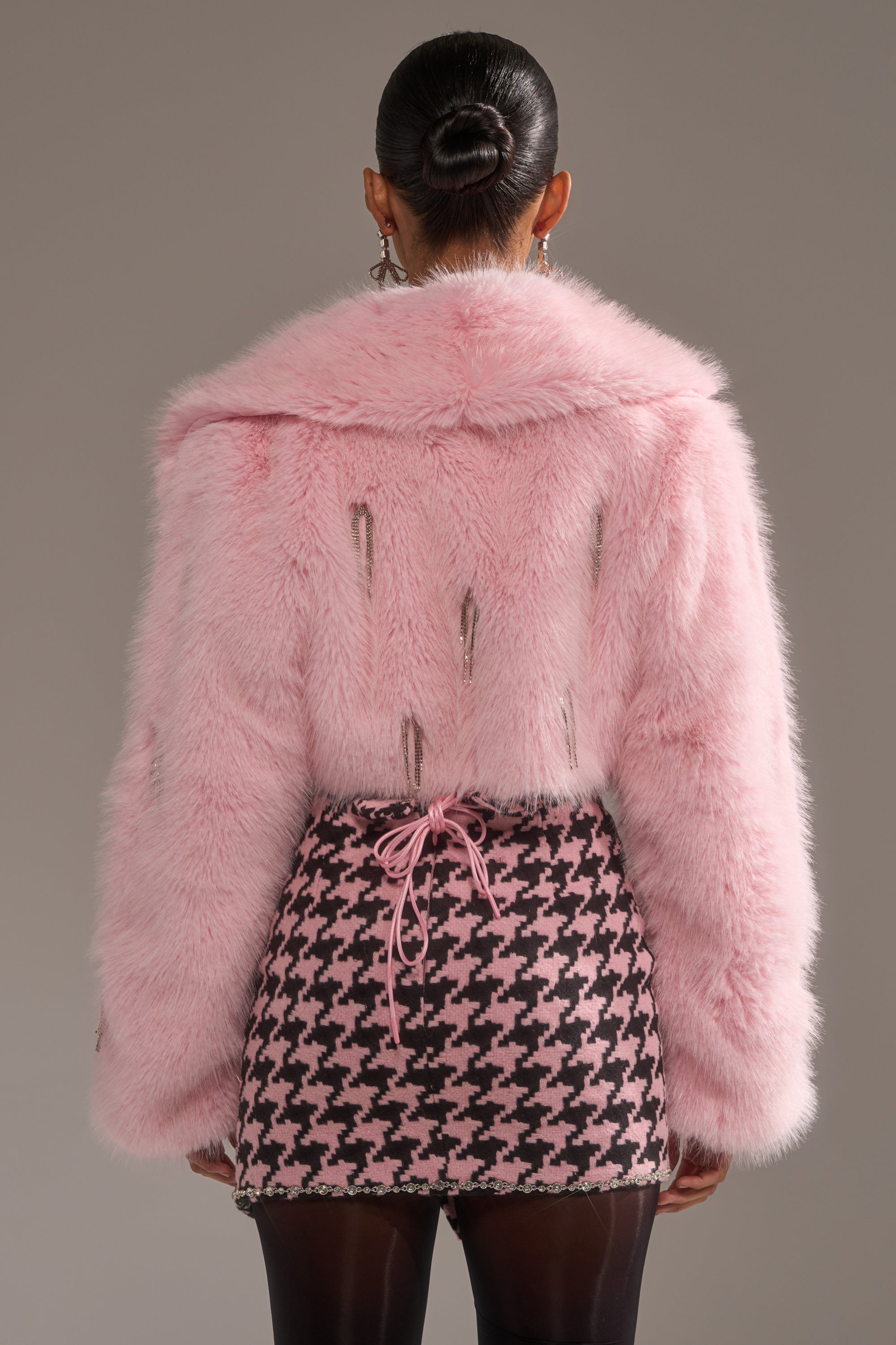 A person with dark hair in a bun wears the SWEET TALK EMBELLISHED FAUX FUR jacket in pastel pink with silver zippers, paired with a pink and black houndstooth lace-up mini skirt, against a gray background. 5