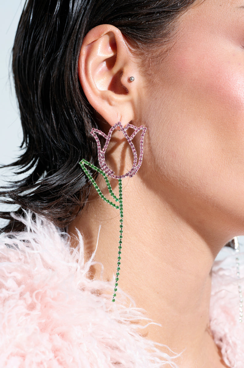 A person wears TULIP TIME EARRING featuring pink petals and a green stem with sparkling rhinestones. Their short dark hair pairs well with the pink fluffy garment, embodying a chic springtime look against the soft light background.