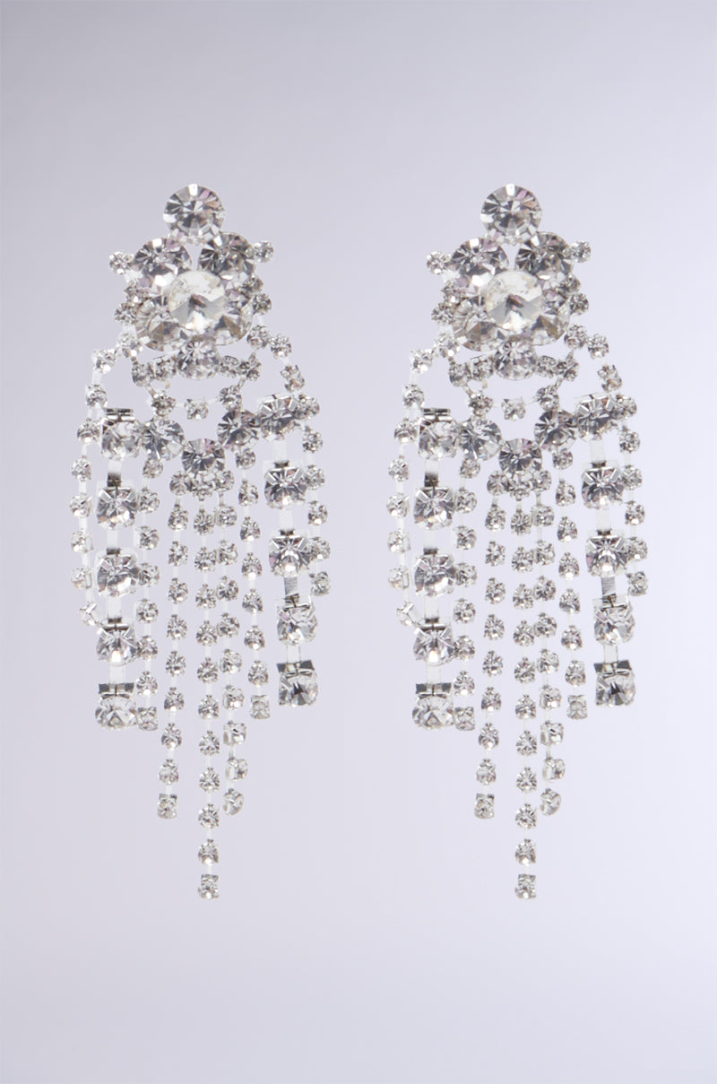 The CALL ME QUEEN EARRING features a dazzling pair of faux silver chandelier earrings embellished with multiple strands of shimmering rhinestone fringe, designed to capture light and offer an elegant, glamorous look against a subtle gray background.