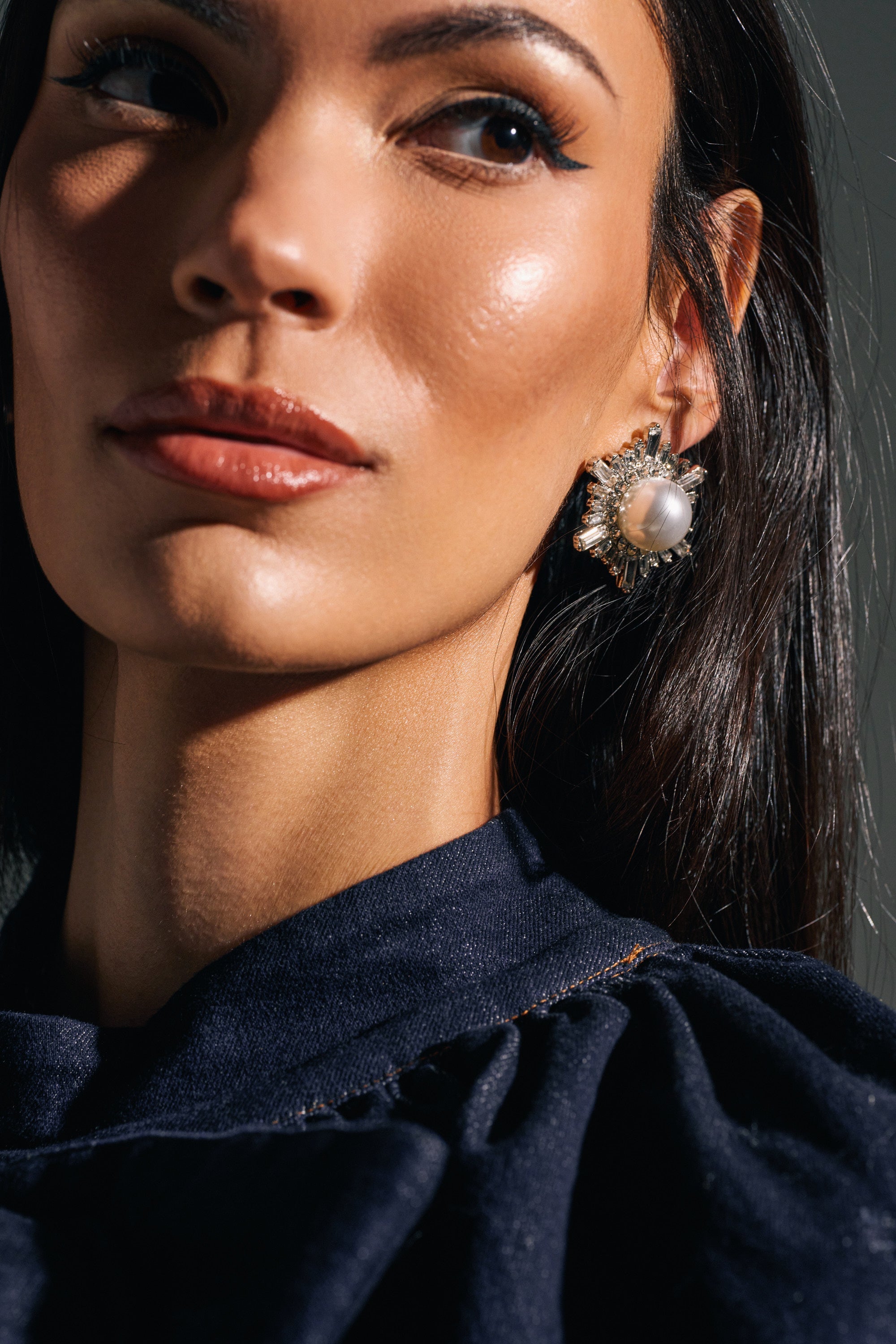 A woman with glowing, smooth skin and winged eyeliner wears the TAY EARRING—oversized pearls with rhinestone detailing. Her straight dark hair and dark navy top highlight this glam accessory, completing her chic look. 2