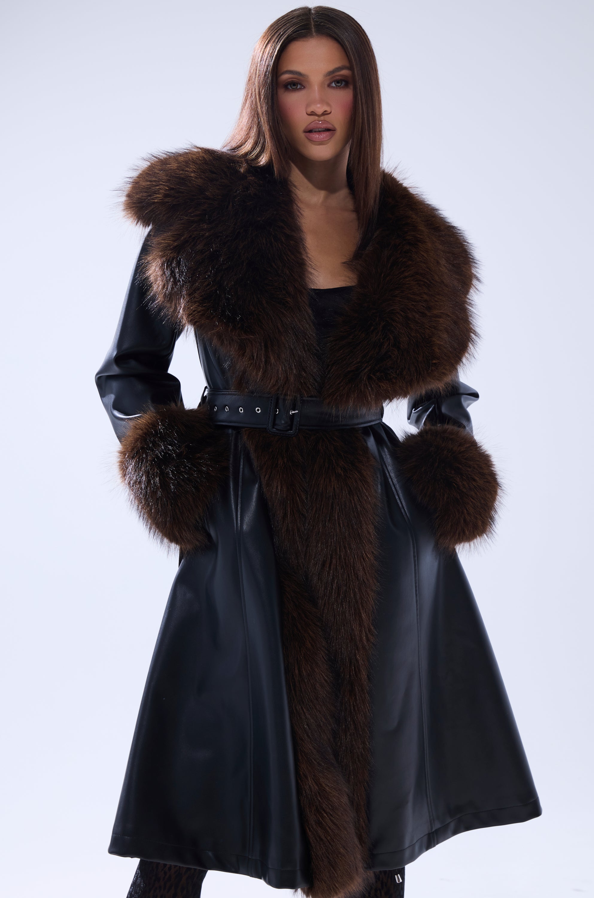 A woman wears the MAIN SQUEEZE FUR TRIM TRENCH, a long black leather coat featuring large brown fur trim on the collar, cuffs, and front. She stands with her hands in her pockets against a plain light background.