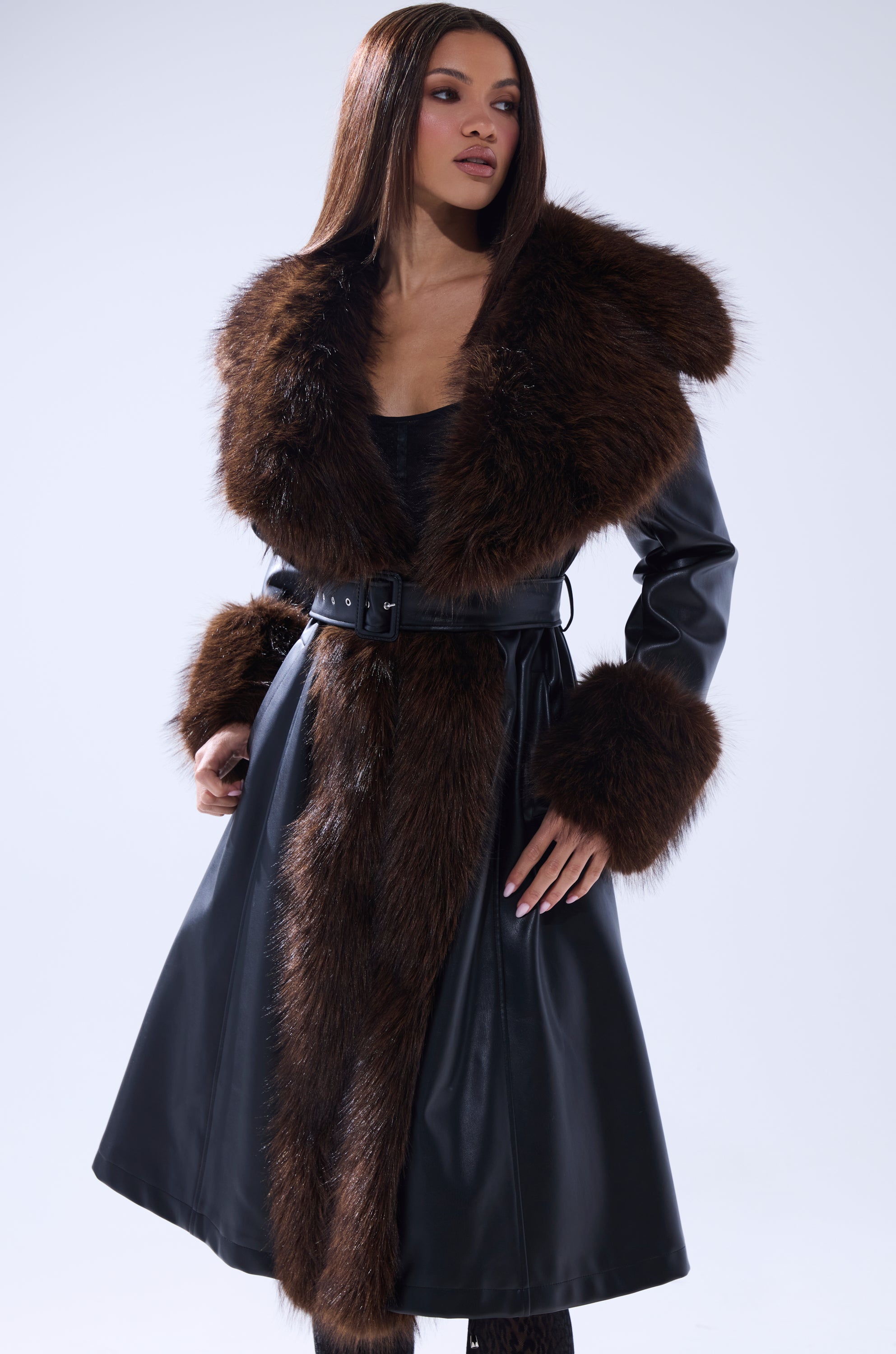 A woman models the MAIN SQUEEZE FUR TRIM TRENCH, a long black leather coat with a wide brown faux fur collar, matching cuffs, and a belted waist, set against a light background.