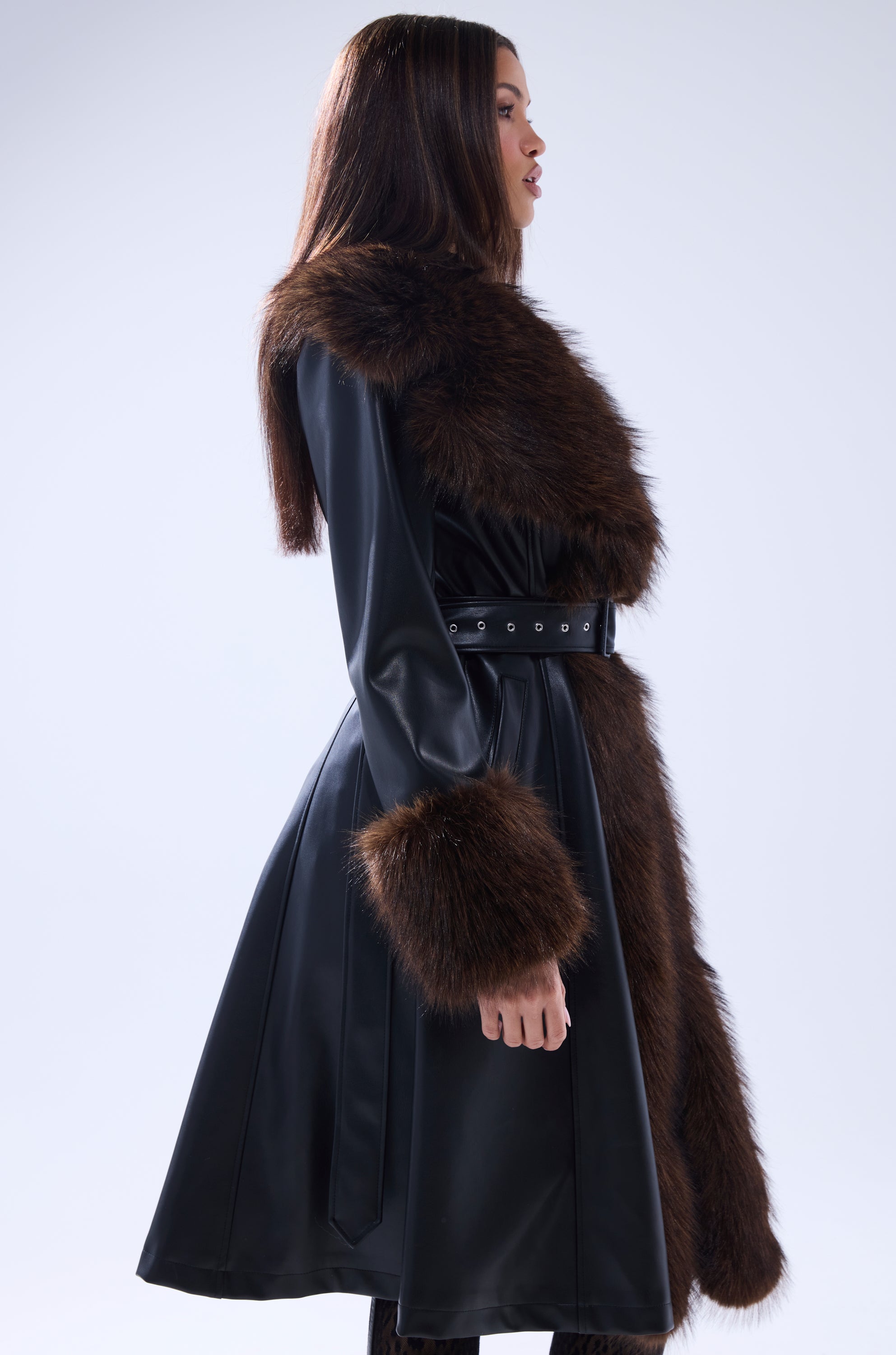 A woman in profile wears the MAIN SQUEEZE FUR TRIM TRENCH, a long black leather coat with a wide brown fur collar and cuffs, belted at the waist, against a light gray background.