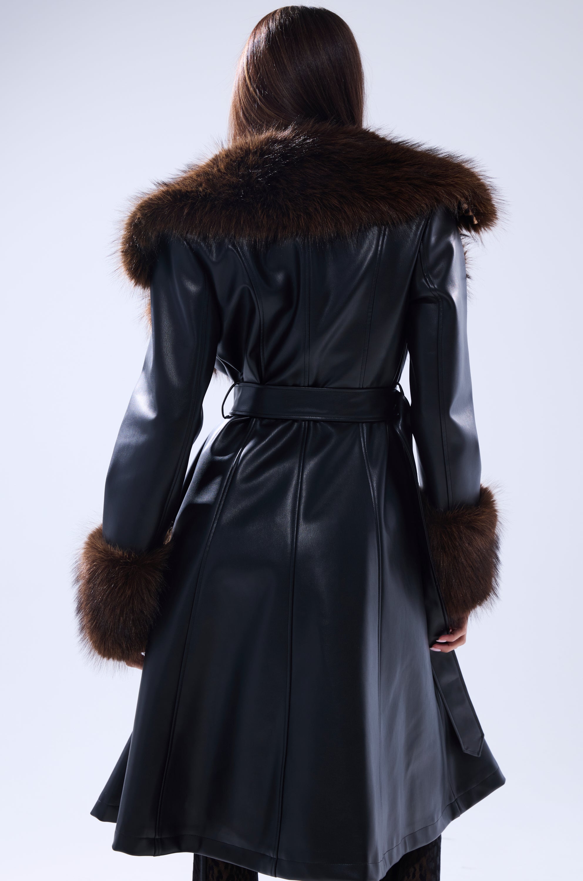 A person wears the MAIN SQUEEZE FUR TRIM TRENCH, a black leather trench coat with a wide dark brown fur collar and matching cuffs, shown from the back. The belted waist and glossy finish complete the look.