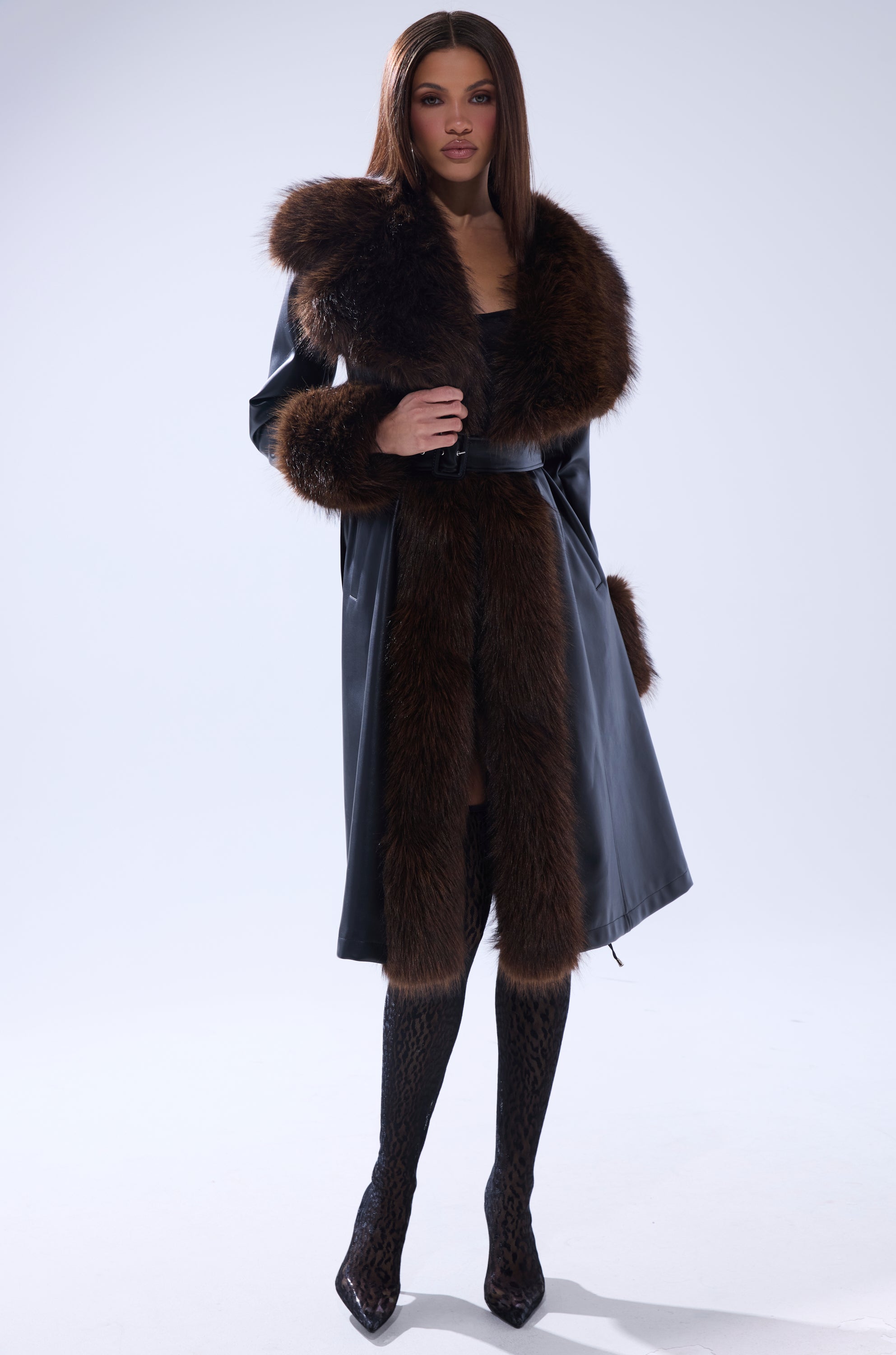 A woman stands confidently in the MAIN SQUEEZE FUR TRIM TRENCH, a long black coat with bold brown fur trim, paired with patterned black stockings and pointed heels against a plain light background.