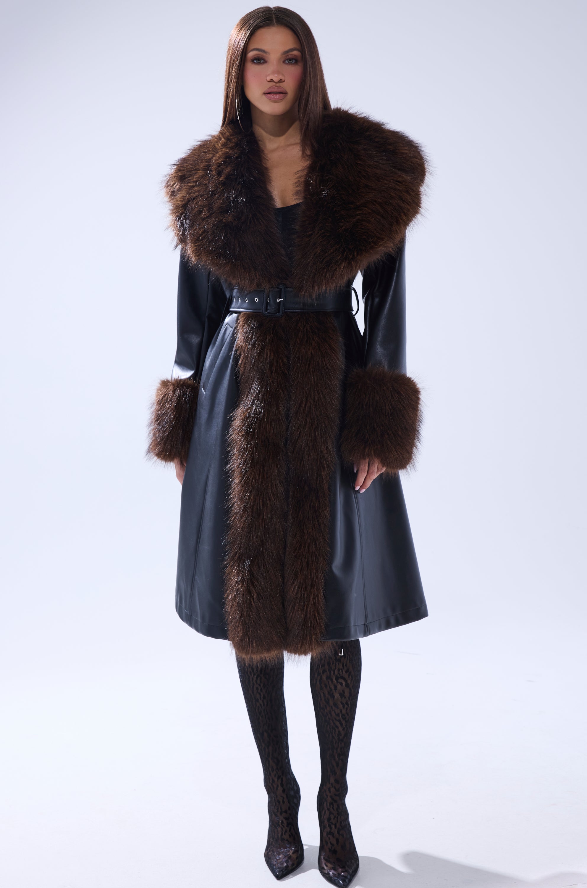 A woman stands facing forward in the MAIN SQUEEZE FUR TRIM TRENCH, a long black leather coat with wide brown fur trim on the collar, cuffs, and front. She pairs it with patterned tights and pointed heels, her straight brown hair worn loose.
