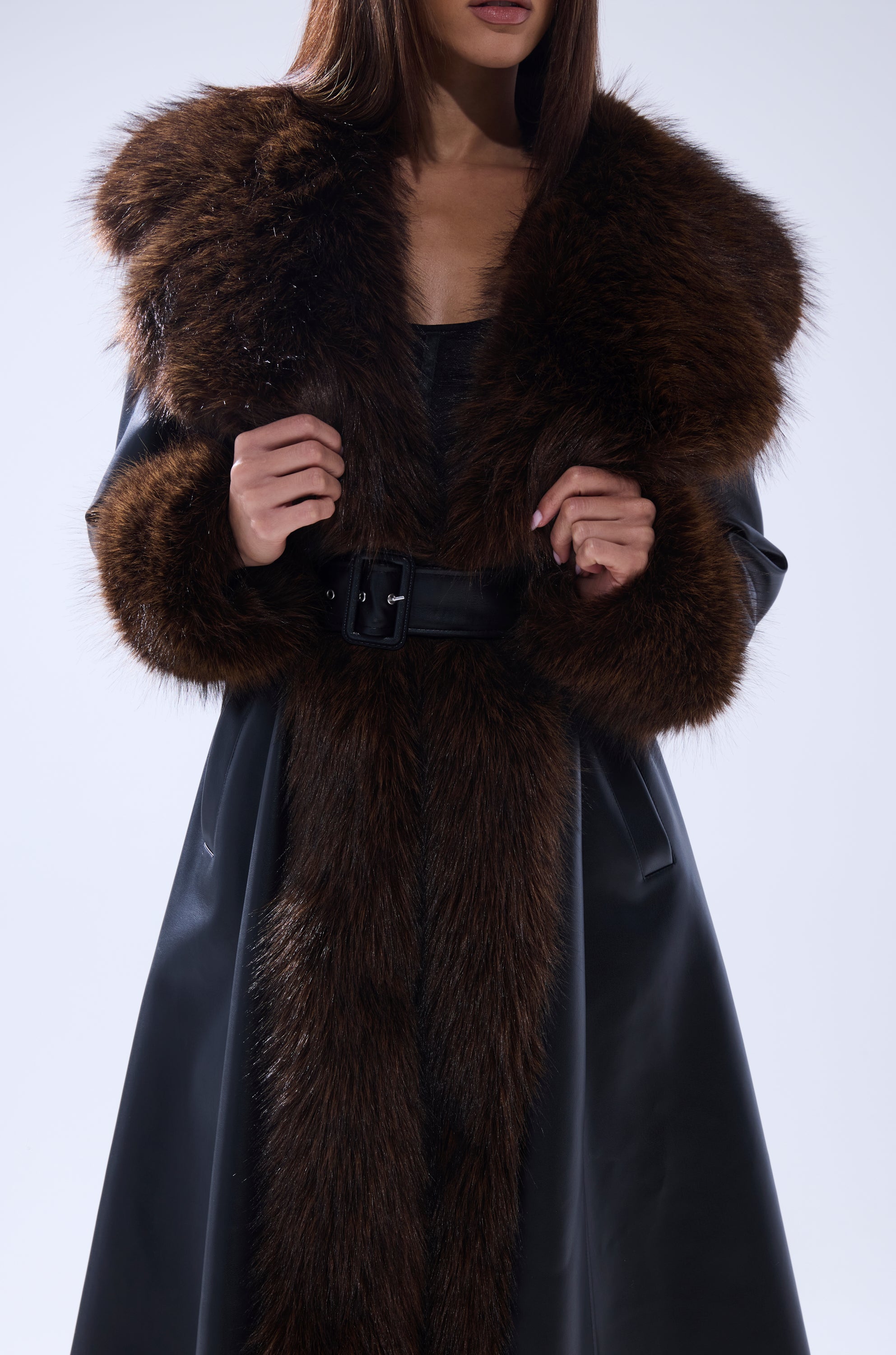 A woman wearing the MAIN SQUEEZE FUR TRIM TRENCH, a black coat with a large dark brown fur collar and cuffs, holds the collar with both hands; her face is partially out of frame.