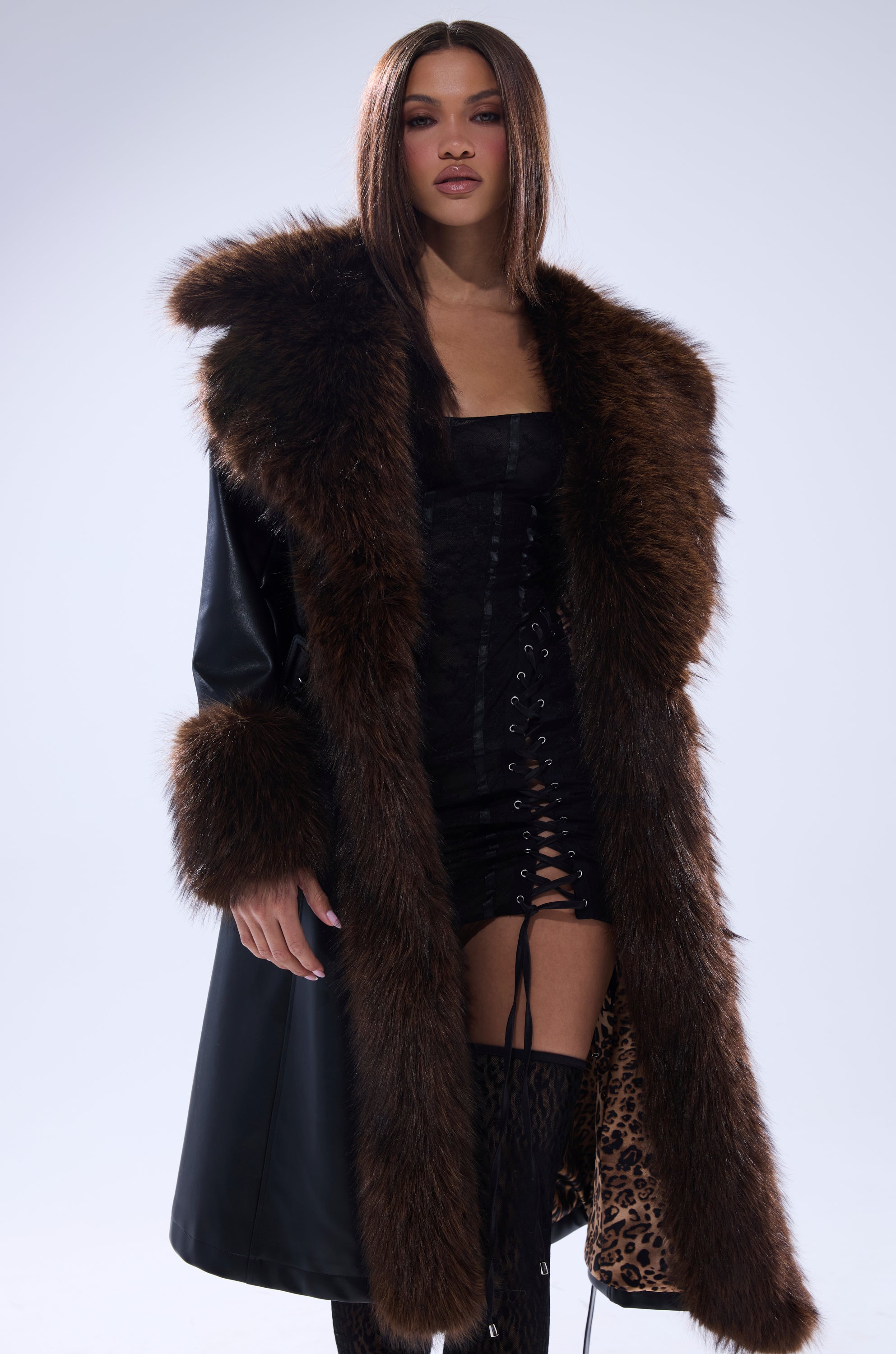 A woman models the MAIN SQUEEZE FUR TRIM TRENCH, a long black coat with large brown fur trim on the collar and cuffs, styled with a black lace-up dress and thigh-high black boots against a plain light background.