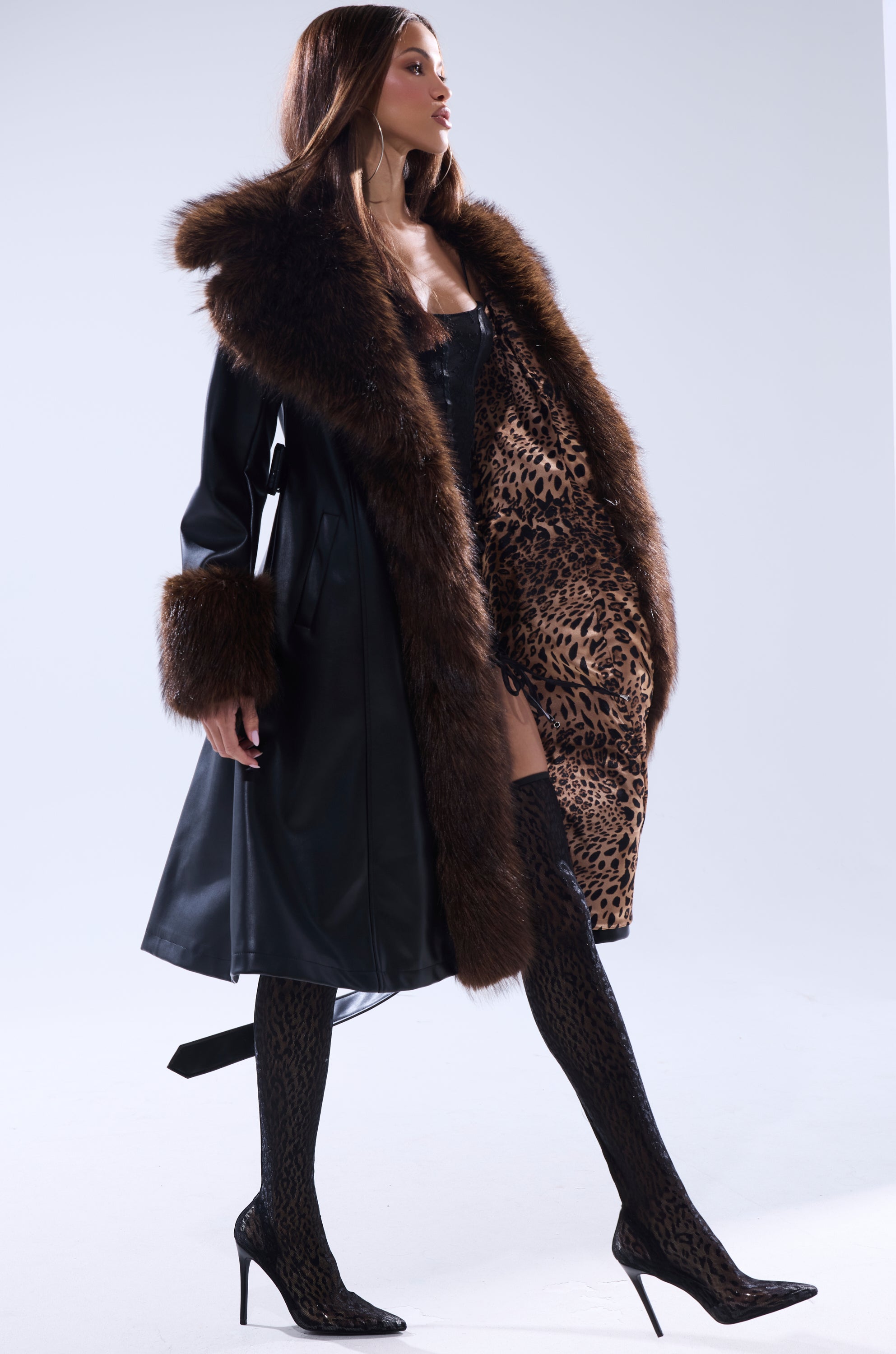 A woman poses confidently in the MAIN SQUEEZE FUR TRIM TRENCH, a black leather coat with a large fur collar, cuffs, and leopard print lining. She pairs it with thigh-high patterned stockings and black heels against a plain light background.